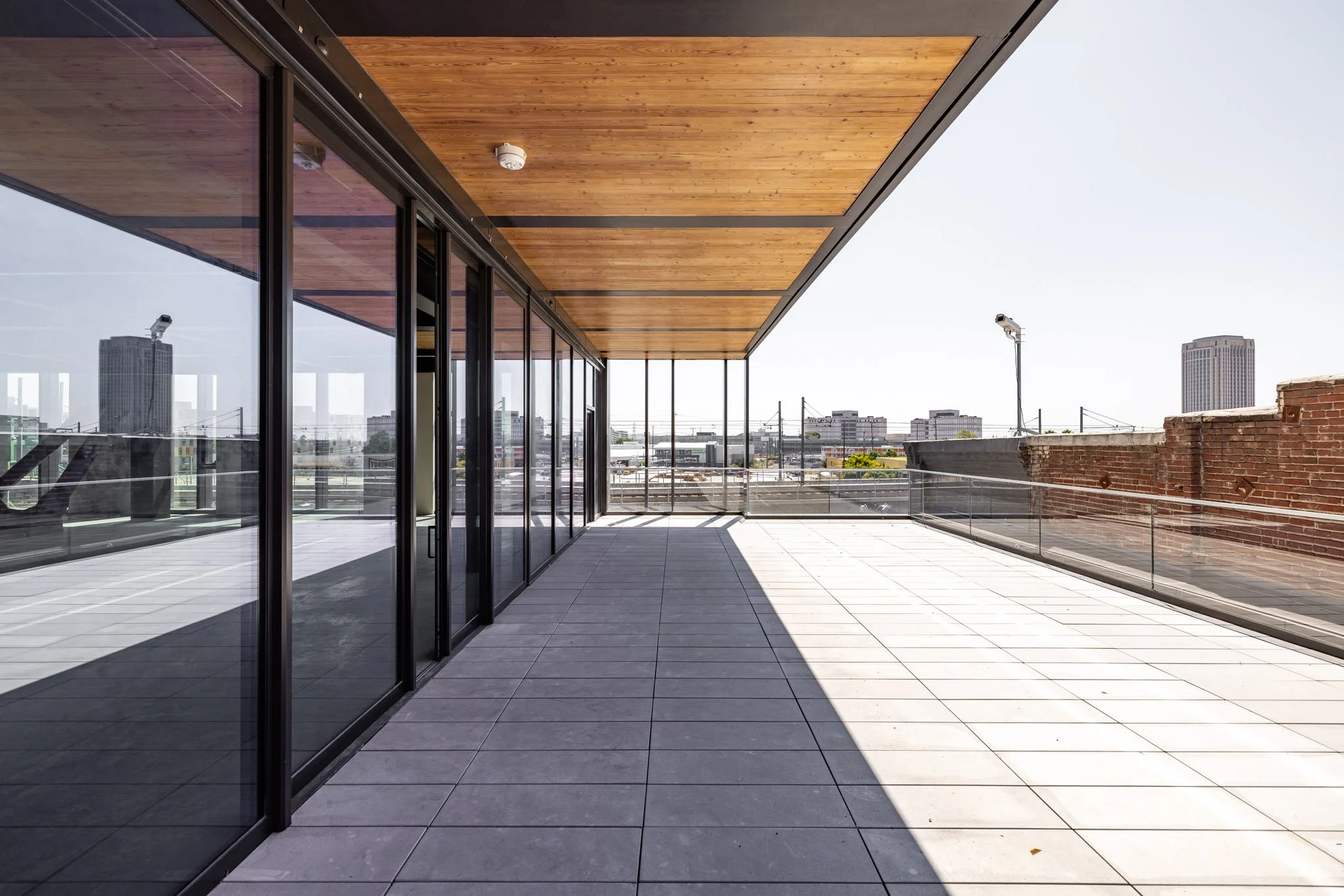 Empty outdoor balcony with glass railings, tiled floor, and a wooden ceiling, overlooking a city skyline with tall buildings.