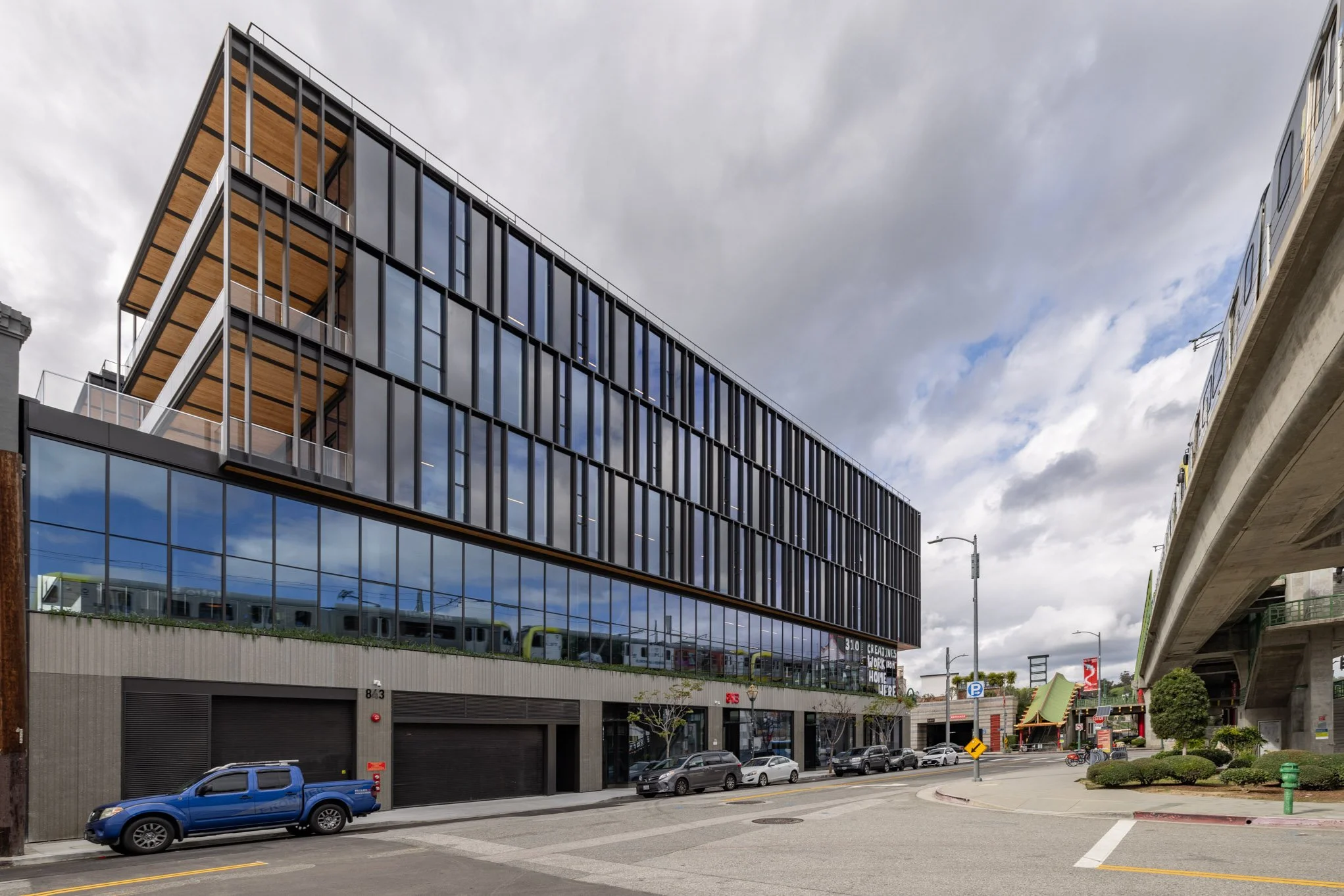 Modern multi-story building with glass windows and a staircase on the exterior, parked cars along the street, and an elevated train track nearby under a cloudy sky.
