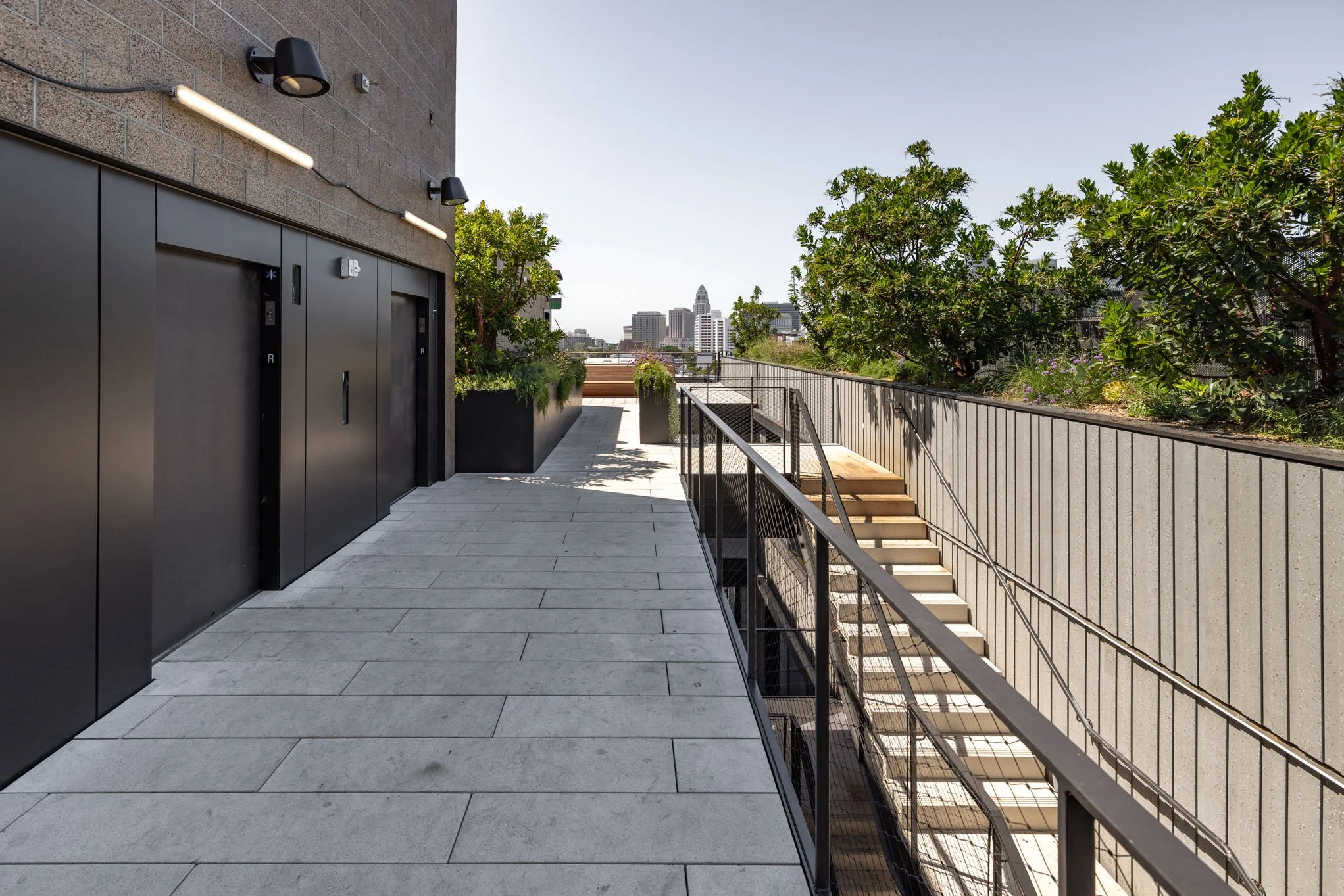 Outdoor terrace with stairs, trees, and city skyline in the background, next to black elevator doors and modern wall-mounted lights.