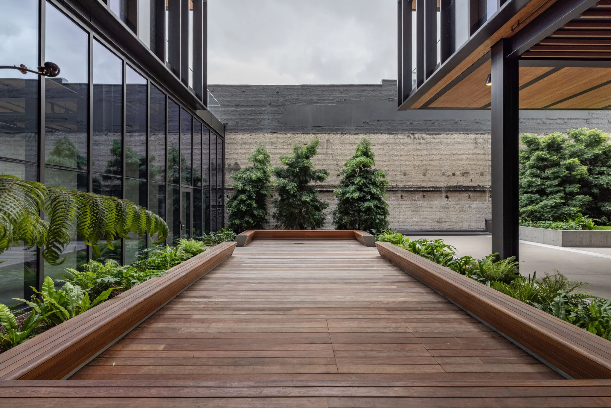 A modern outdoor patio area featuring wooden decking, surrounded by greenery and trees, with glass walls on one side and steel supports on the other, under a cloudy sky.
