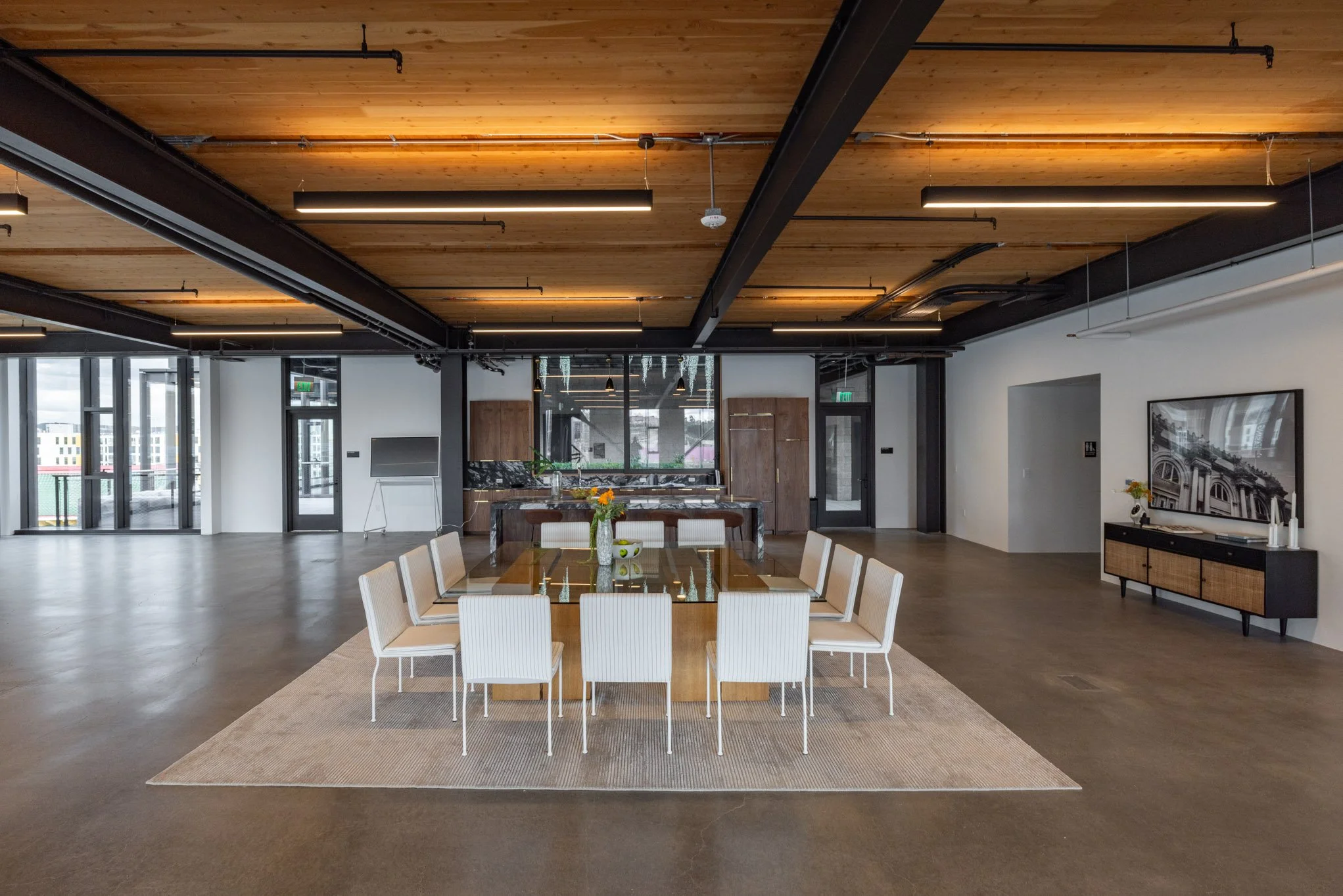 Modern open-concept dining area with a large glass-topped dining table surrounded by white chairs, beige rug, serving buffet, and a living space with a TV on a black sideboard, natural light from glass doors, and a kitchen with dark wood cabinets and