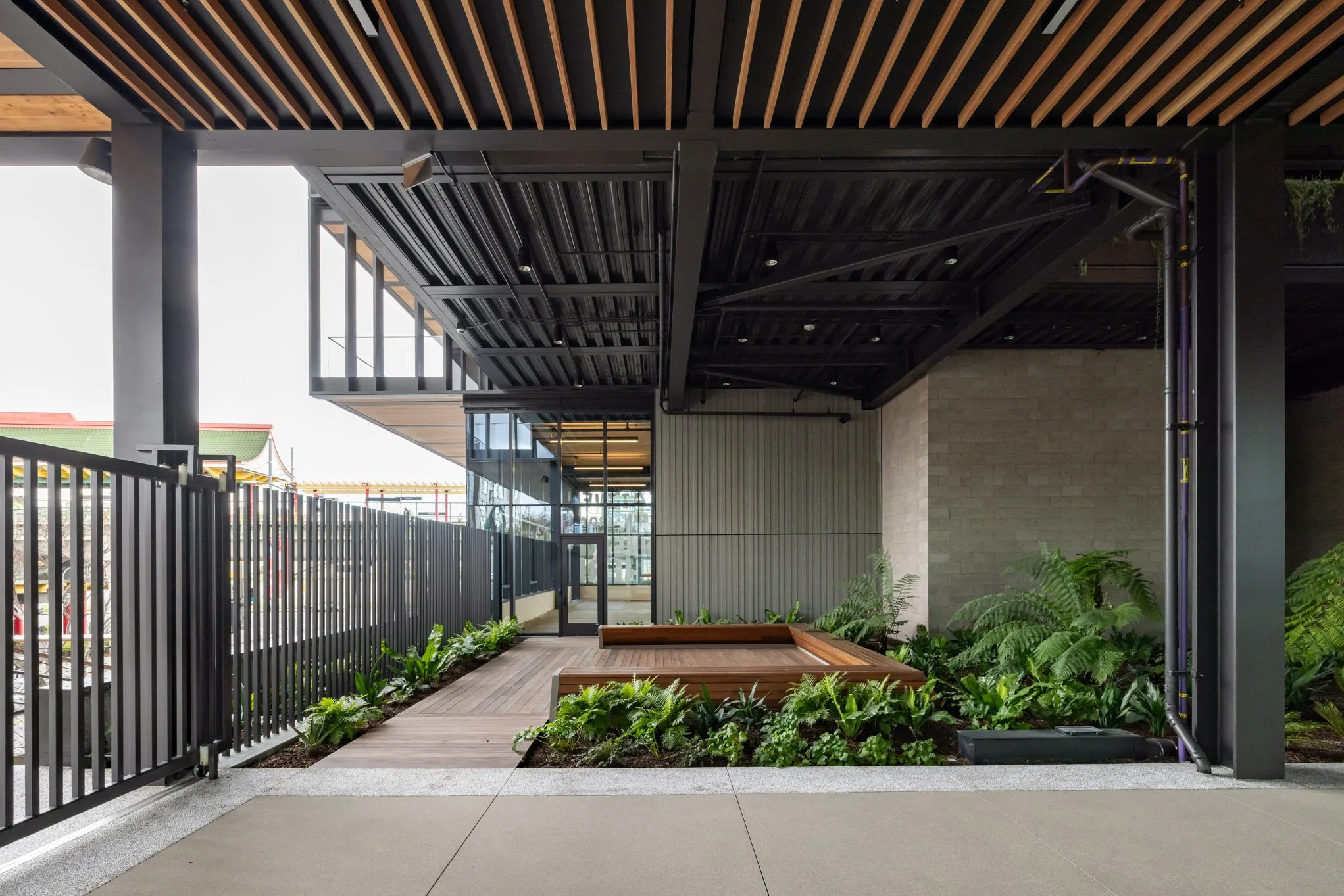 Modern building exterior with metal and glass construction, wooden accents, a landscaped garden bed, and a wooden bench, featuring a black metal fence and pipes along the exterior wall.
