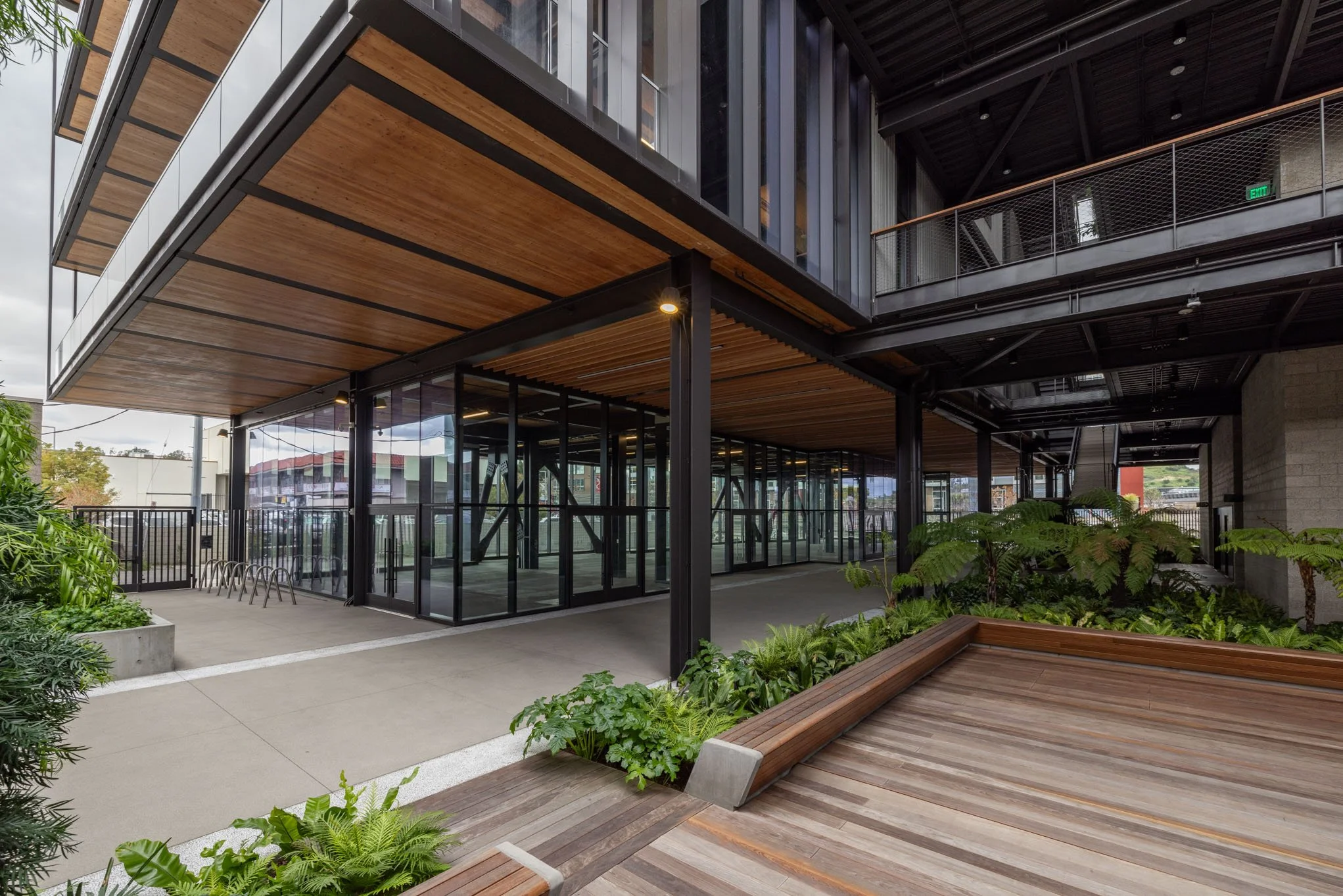 Modern multi-level building with glass walls, wooden overhangs, outdoor seating area, and lush greenery.