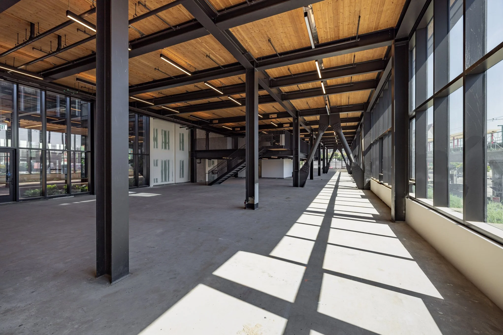 Empty commercial or office space with large windows, exposed wooden ceiling, black metal beams, and staircase, sunlight casting shadows on the floor.