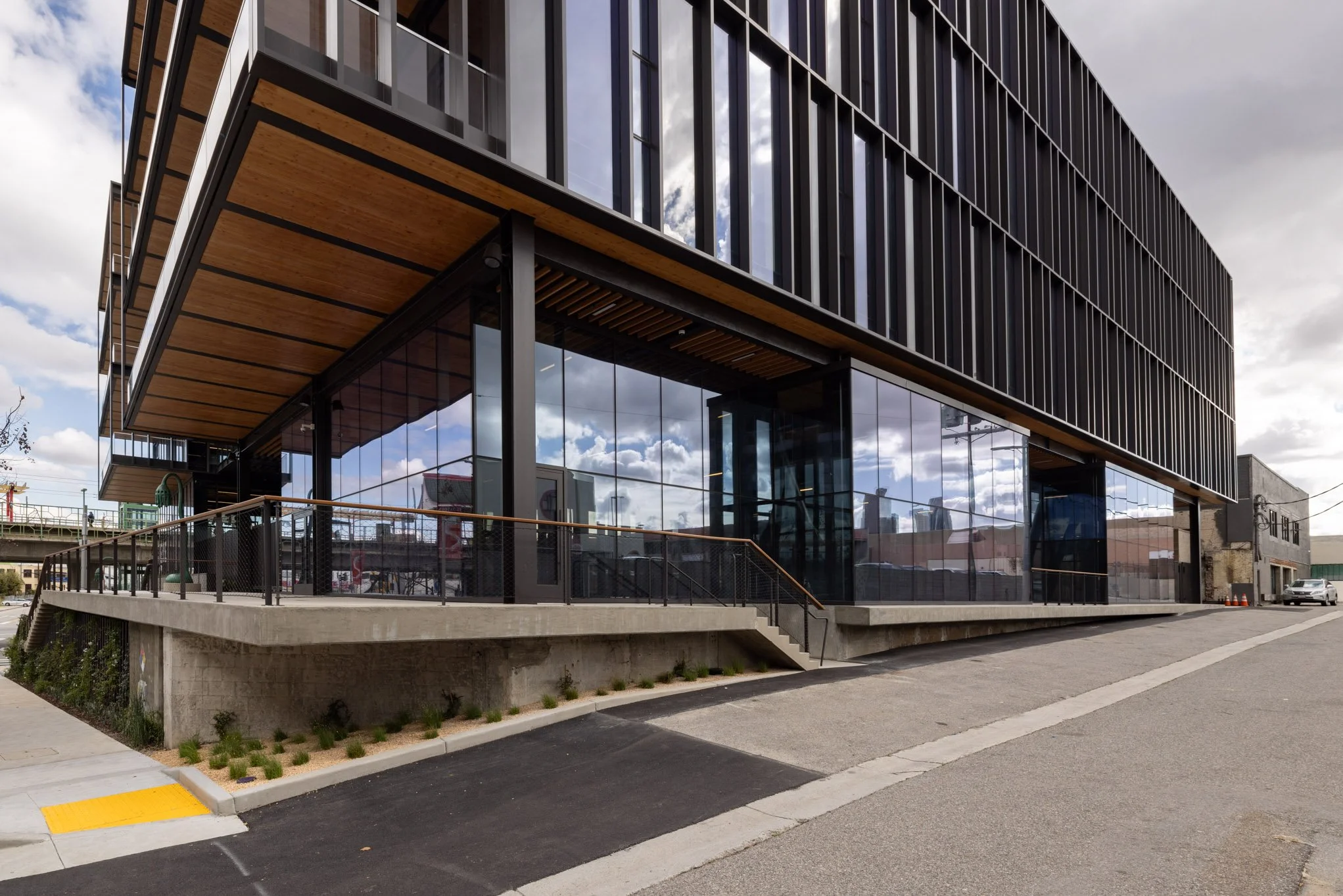 Modern building with glass windows, black metal siding, and wooden accents, located on a sloped street with parking area and sidewalk.