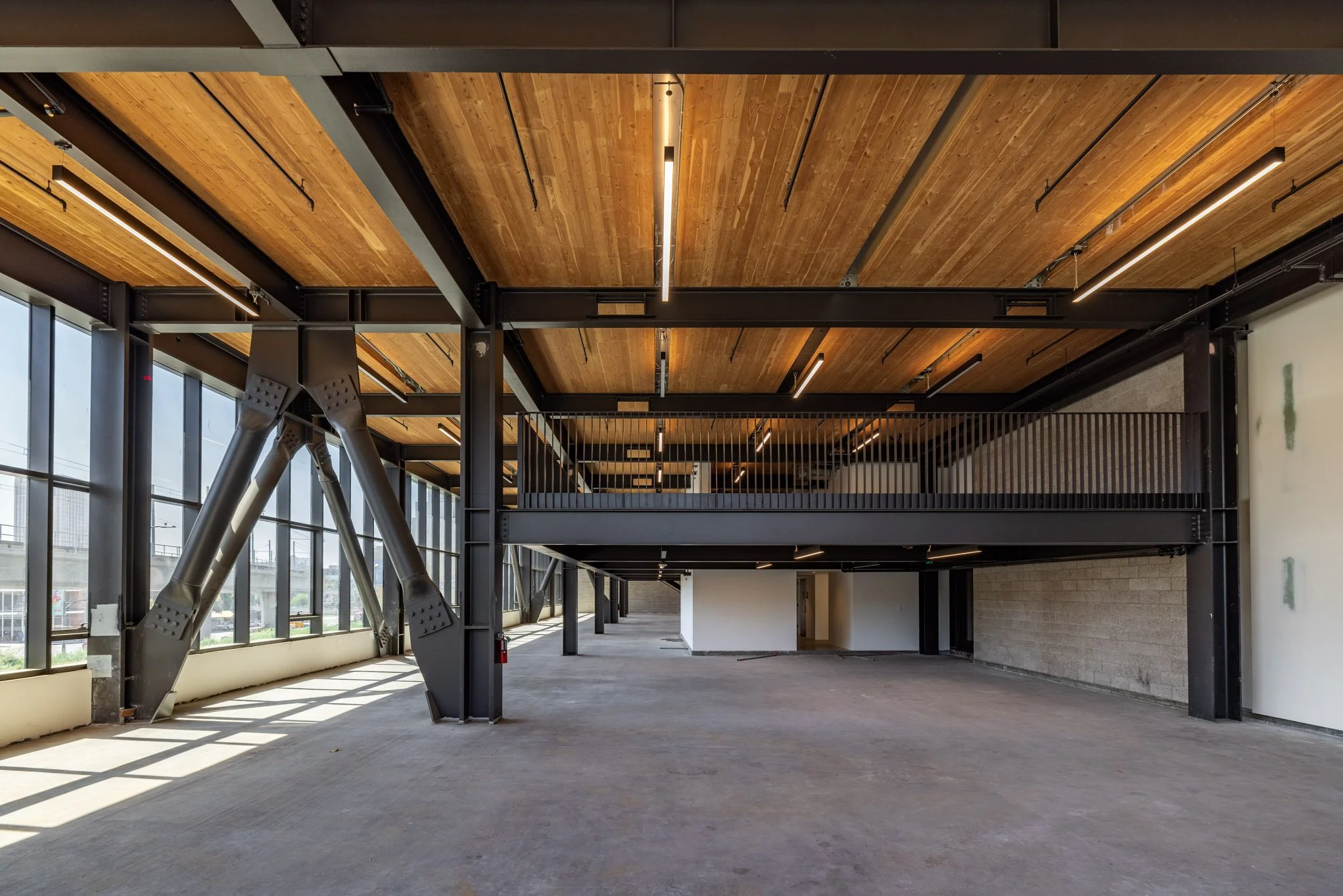 Empty industrial-style interior space with large windows, exposed steel beams, wood ceiling, and a two-story mezzanine with black railing.