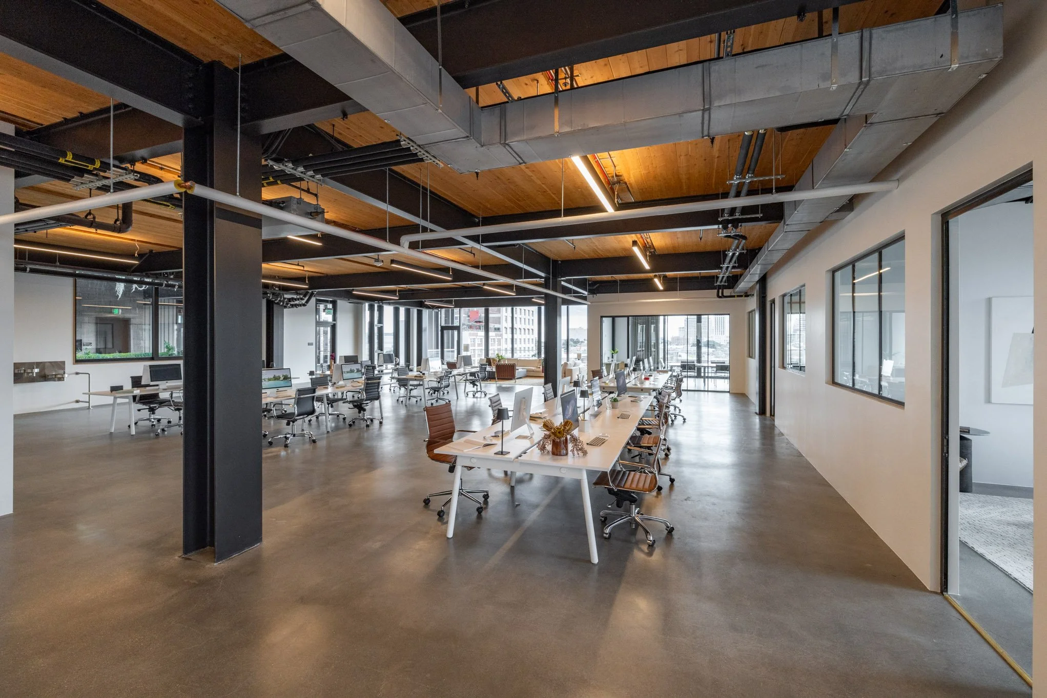 Modern open-plan office space with white desks, black chairs, large windows, and exposed ceiling with wooden panels and industrial ducts.
