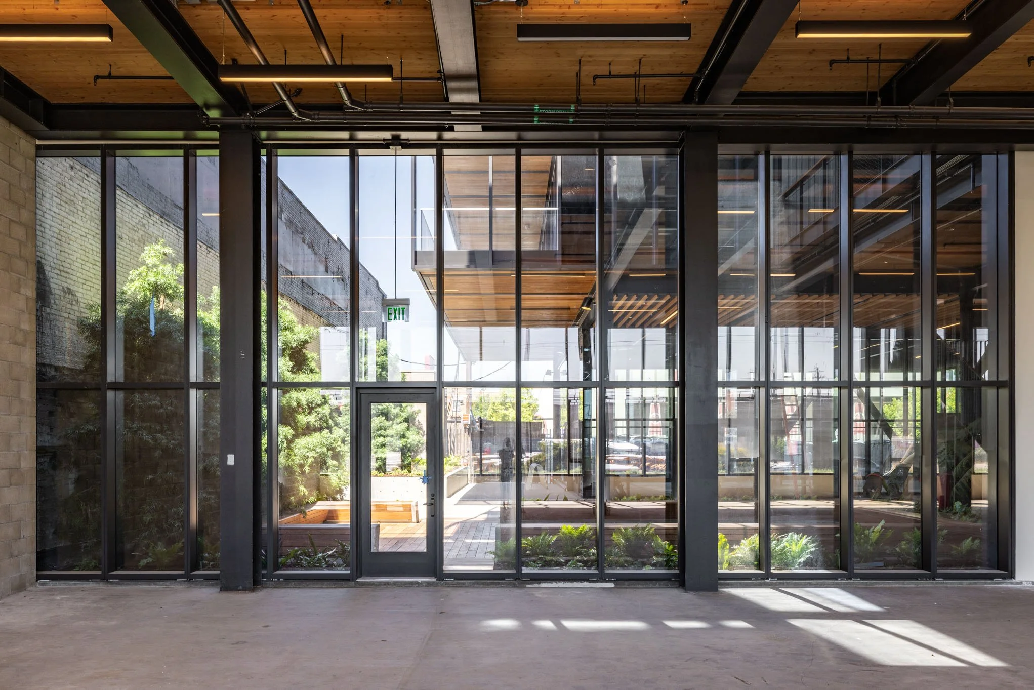 Modern building entrance with large glass windows and door, view of an outdoor patio and greenery, with a staircase and urban surroundings visible outside.
