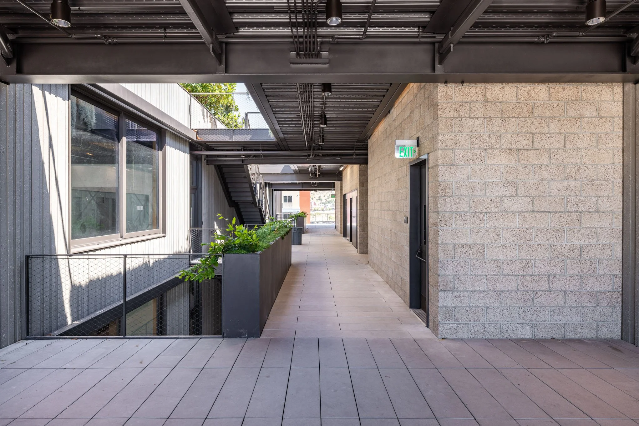 Modern outdoor corridor with tiled flooring, planters with green plants, metal staircase, glass windows, and brick and metal walls, illuminated by natural light.