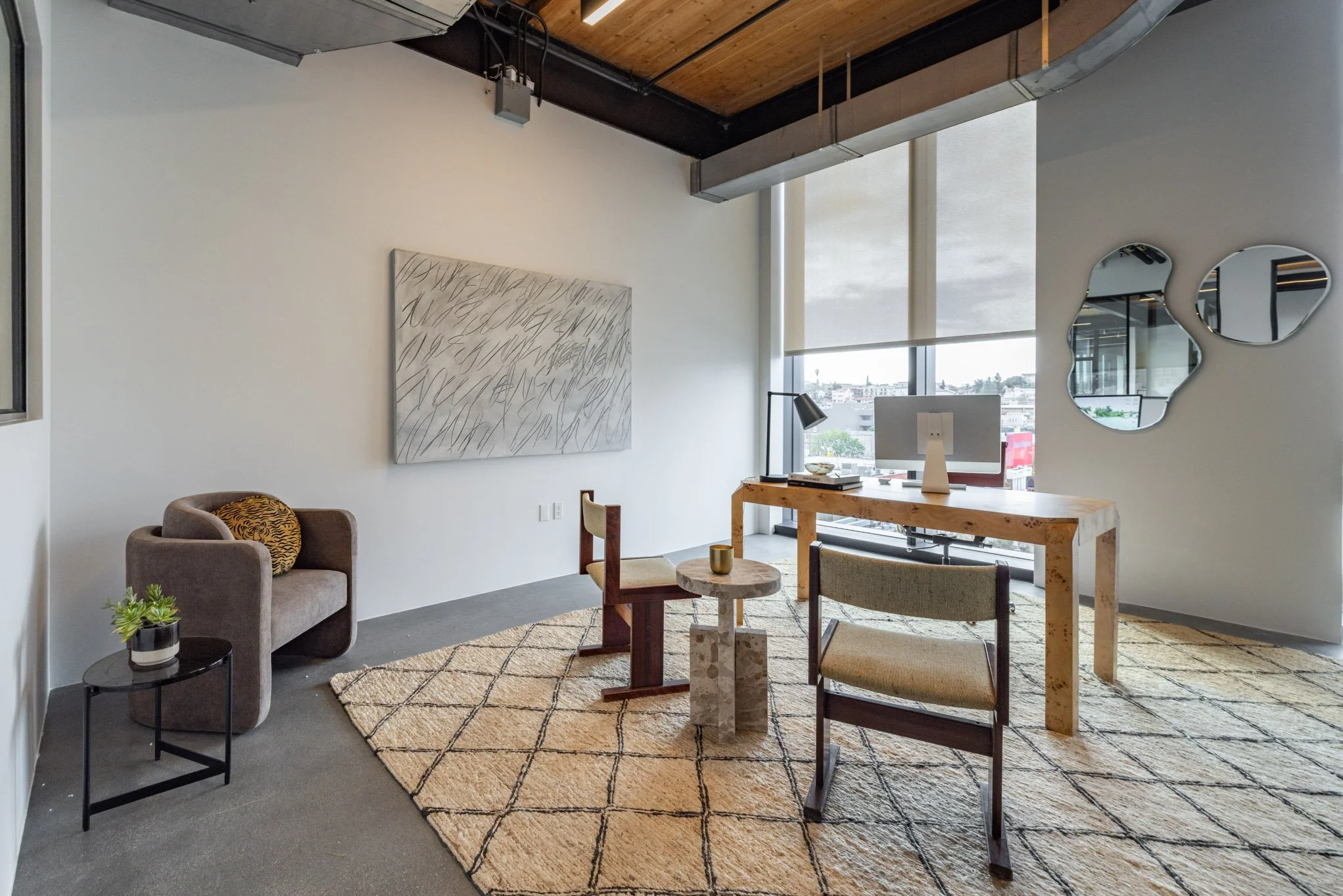 Modern office with a desk, chairs, a computer, wall art, round mirrors, and large windows with city view.