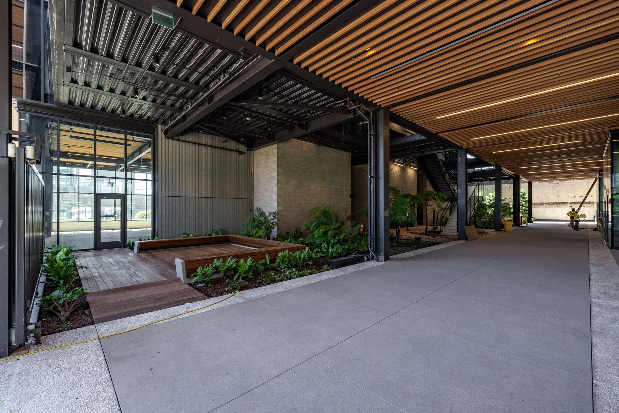 Modern architectural exterior with a covered walkway, black metal staircase, wooden ceiling, glass doors, and landscaped plants.