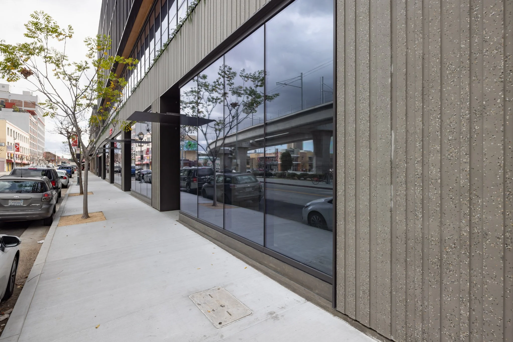 Sidewalk with parked cars and modern building with large reflective glass windows, overcast sky, and a few trees.