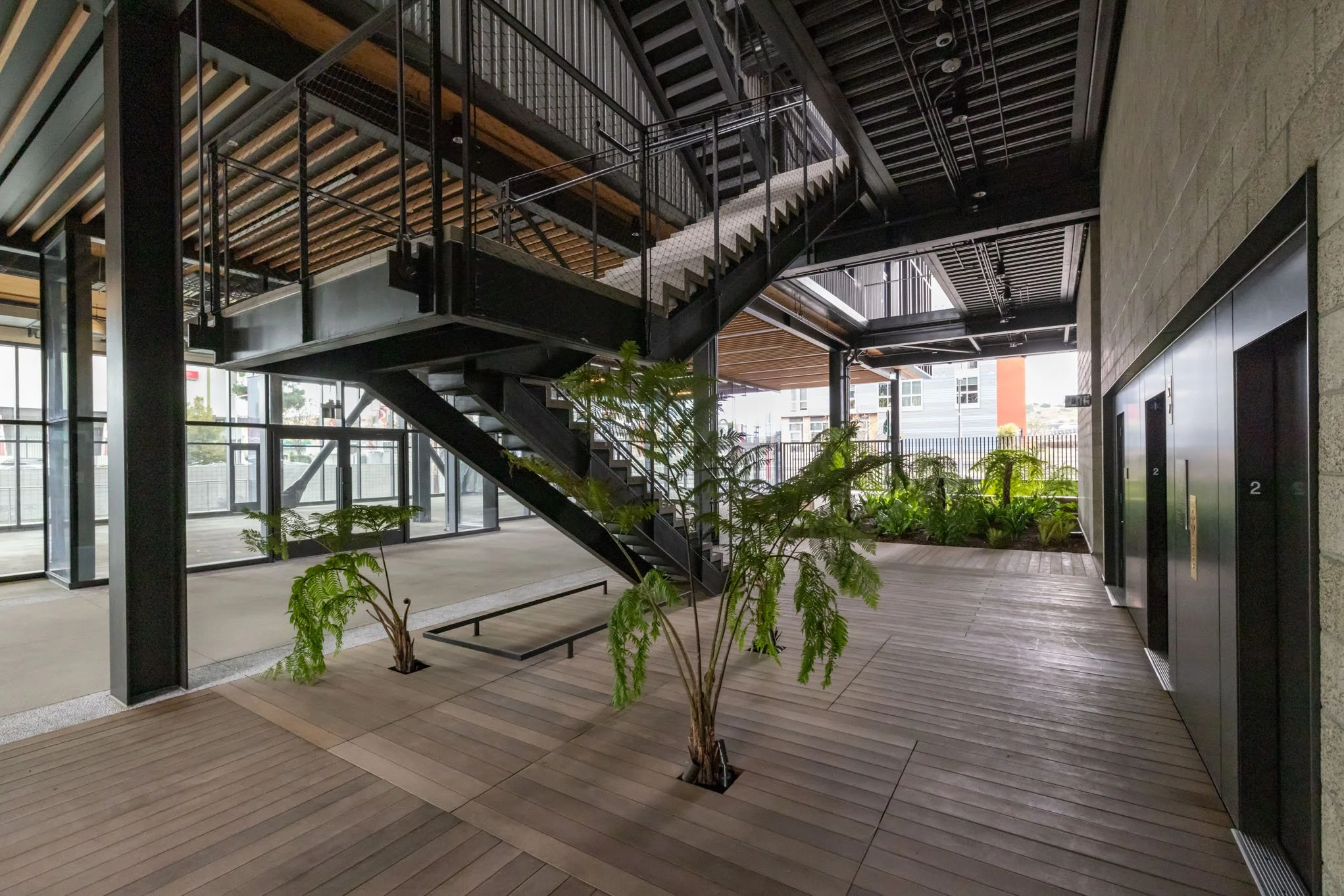 Modern indoor space with a metal staircase, wooden flooring, large windows, two elevators, and plants.