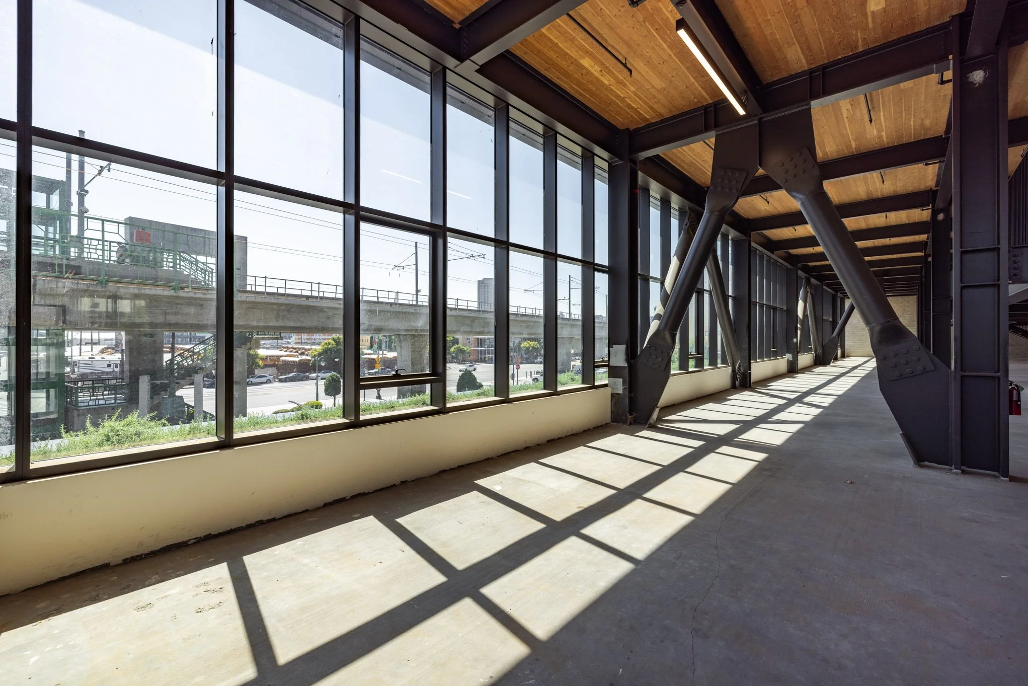 Empty interior space with large floor-to-ceiling windows and sunlight casting shadows, showing an urban view outside and industrial-style black structural supports.