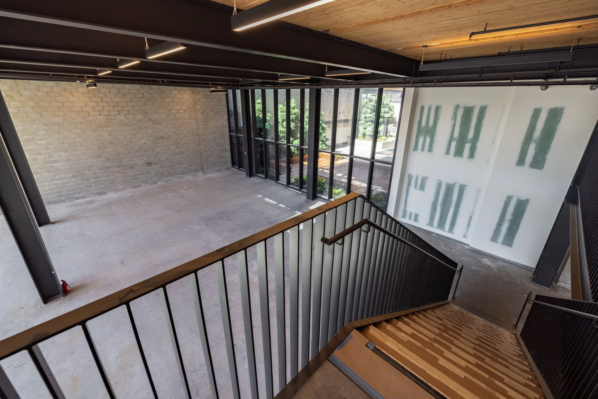 Empty interior space with a staircase, large windows, exposed brick and concrete walls, and unfinished drywall.