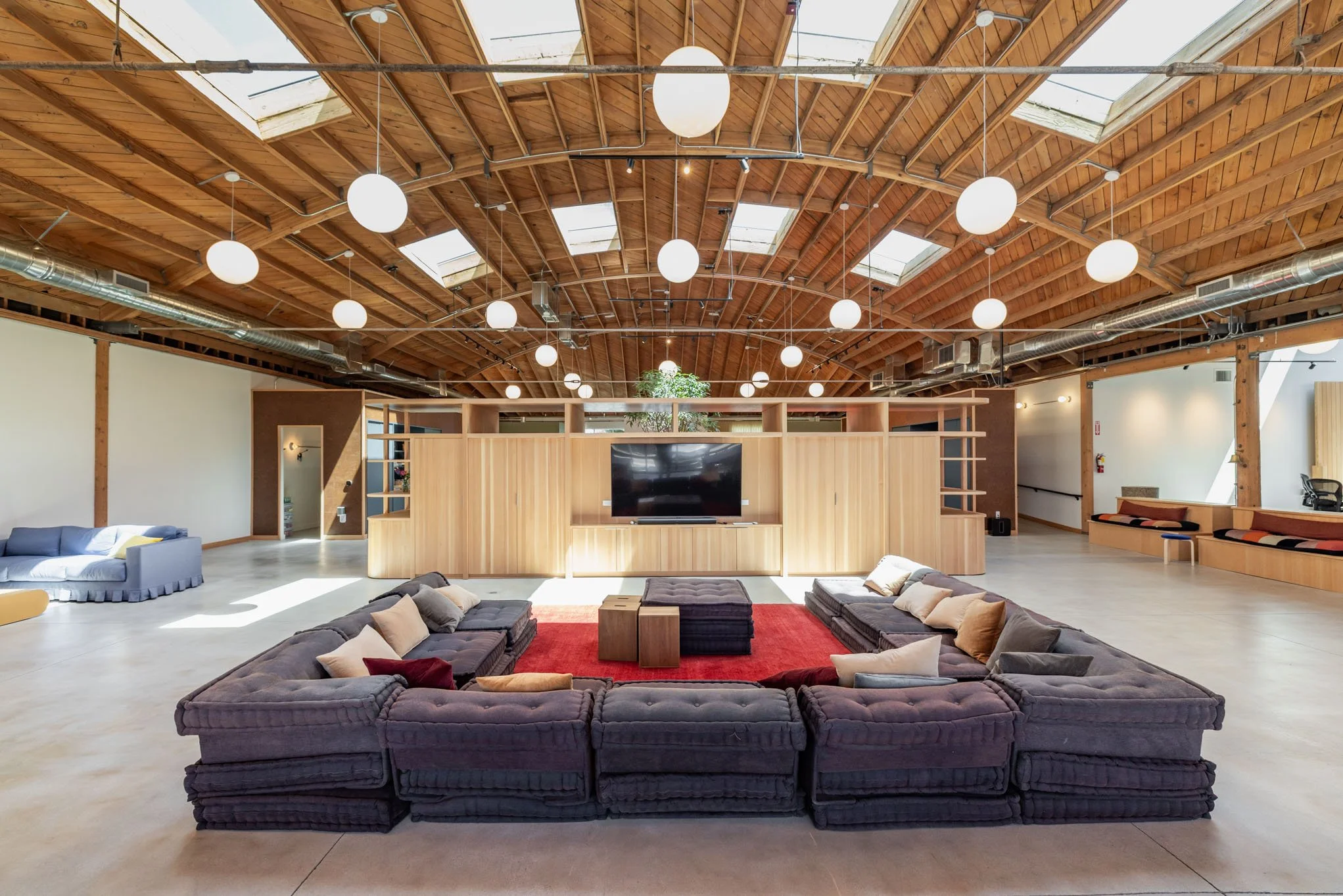 Spacious, modern lounge area with a large sectional sofa, ottomans, and a big flat-screen TV, featuring a high wooden ceiling with skylights and round hanging lights.