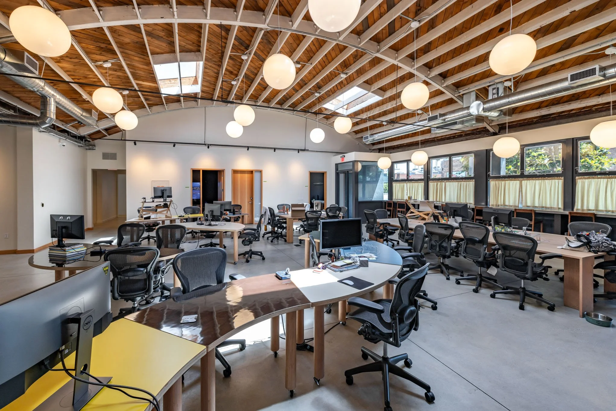 Open office space with multiple desks, ergonomic chairs, desktop computers, and large windows. The ceiling is wooden with exposed beams and round hanging lights.