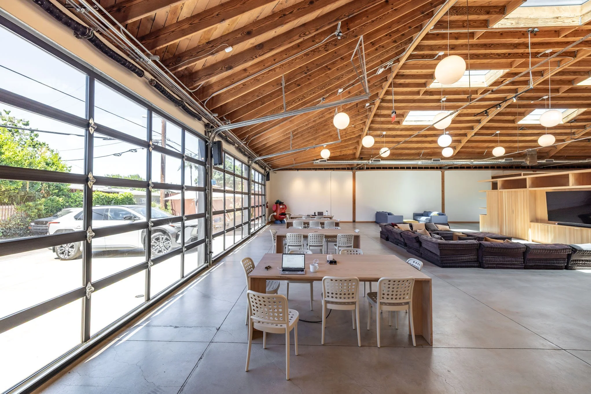 Spacious lounge and meeting area with large garage-style glass doors, wooden vaulted ceiling with hanging round light fixtures, and a mix of tables and couches for seating.