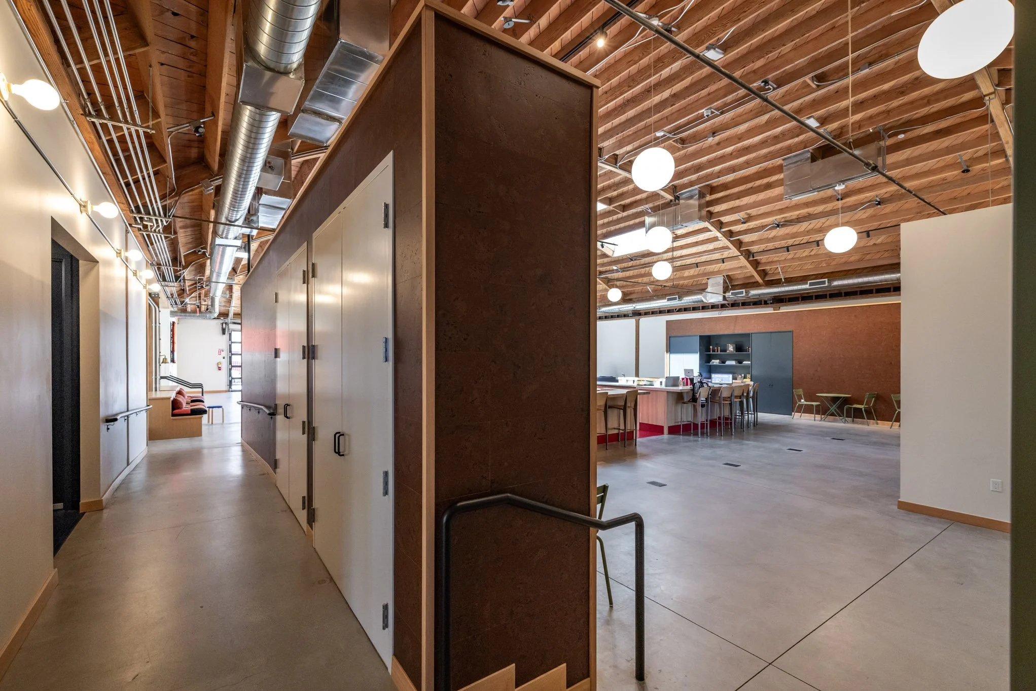 Modern interior space with high wooden ceiling beams, large round white ceiling lights, a curved hallway on the left, a bar or counter area with barstools, and some seating areas with chairs toward the back.