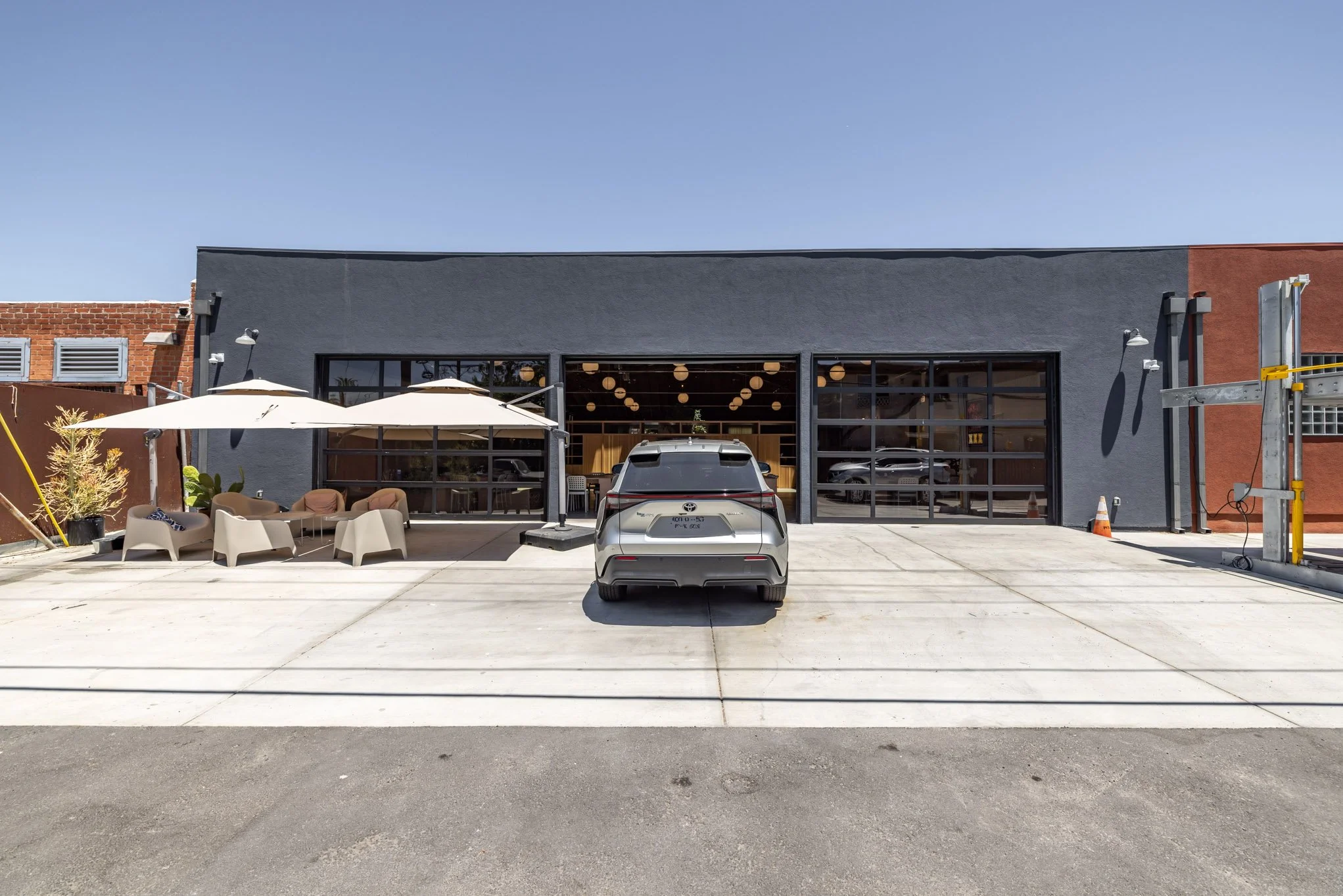 Front view of a modern building with large glass windows, outdoor seating with umbrellas, a parked silver car, and construction materials in the parking lot.