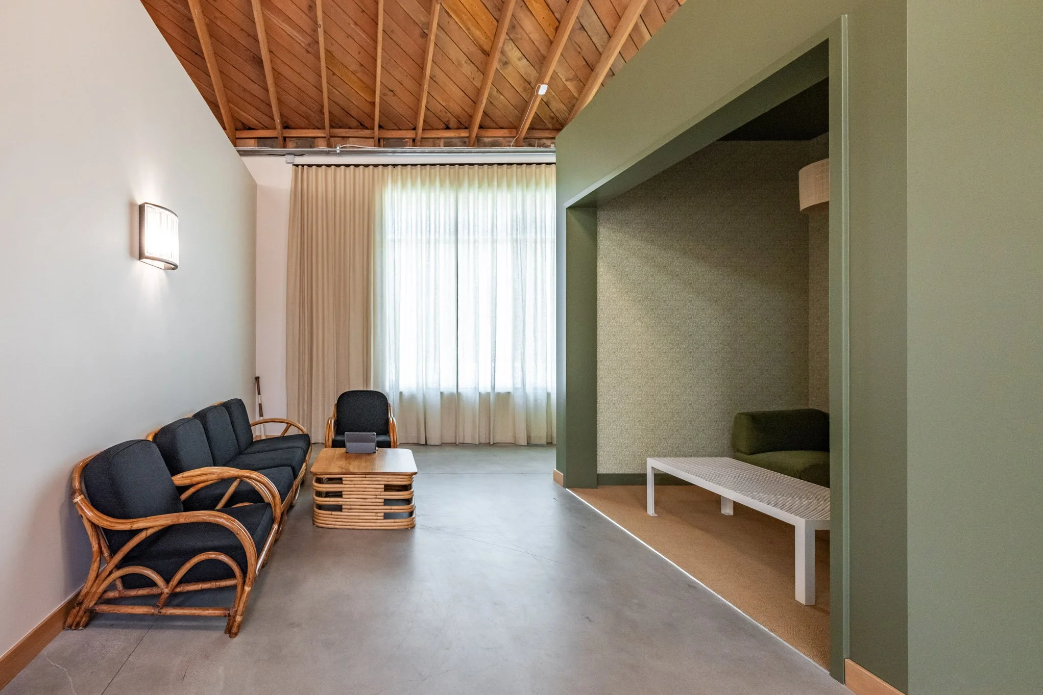 A modern waiting or sitting area with black cushioned chairs with wooden armrests along the left wall, a small wooden coffee table, a black swivel chair on the right, a window with sheer curtains in the background, and a small green alcove with a ben