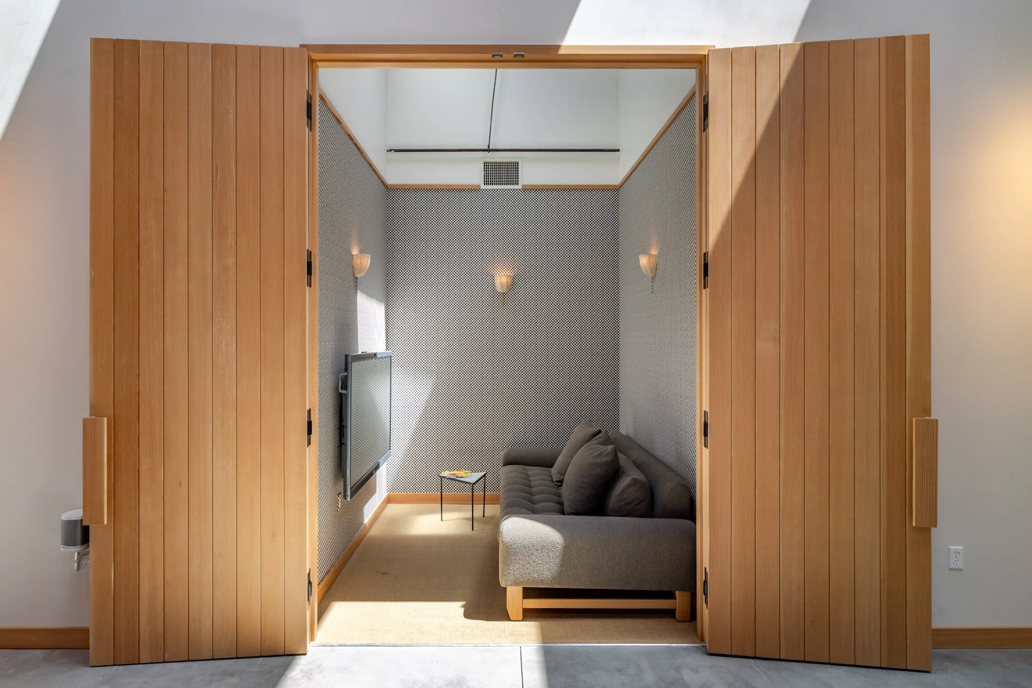 Small enclosed room with a grey sofa, a small black side table, and wall-mounted light fixtures, seen through wooden sliding doors.