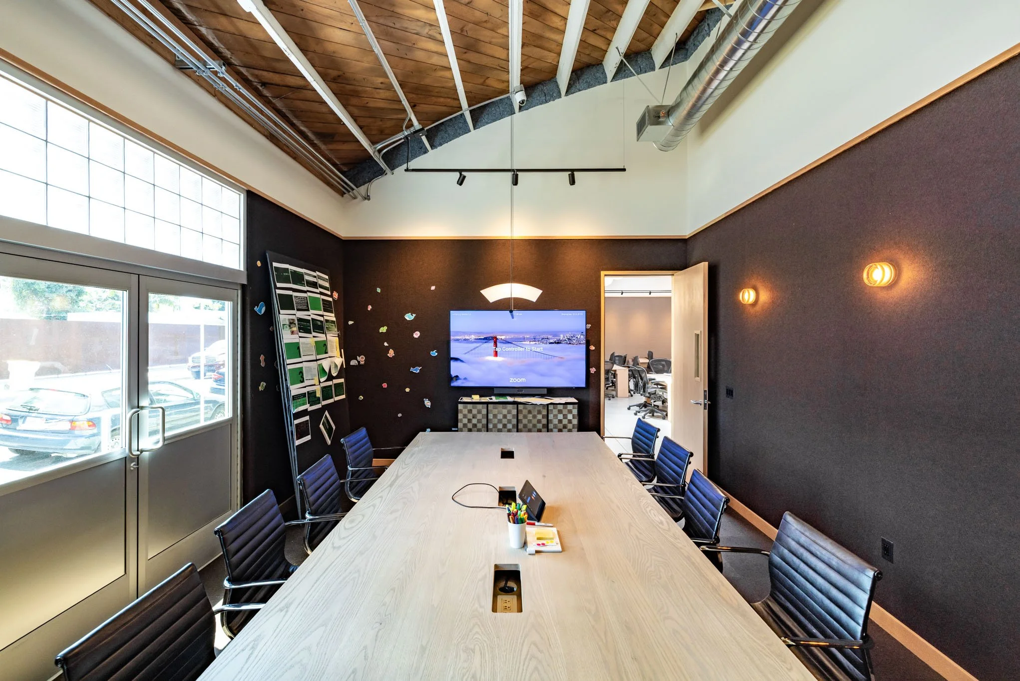 Modern conference room with a long wooden table and black chairs. A large window and door on one side, a wall-mounted screen at the far end displays a screen with the words "Control to start" and a snowy landscape.
