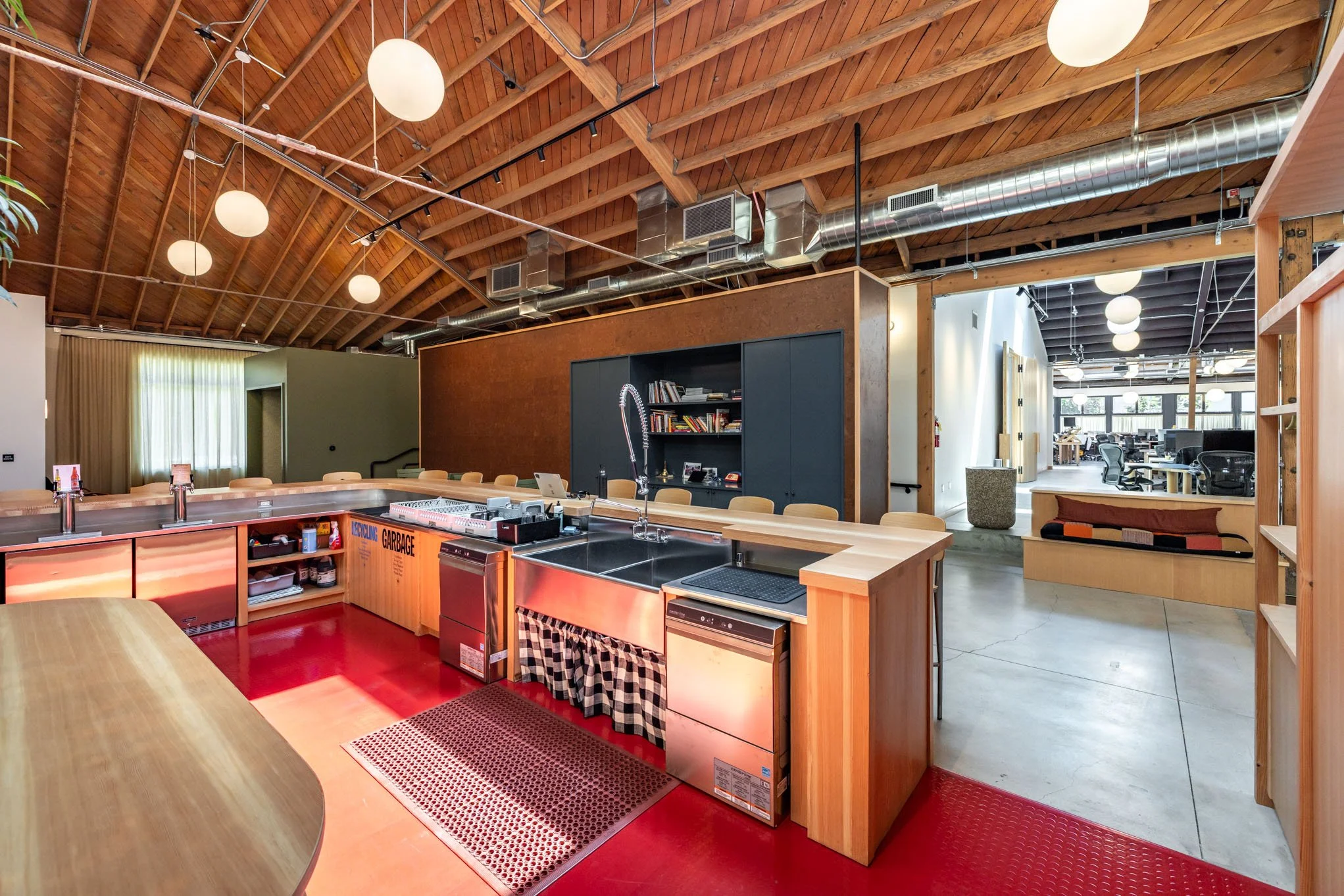 Open-plan kitchen and office space with wooden ceiling and modern decor, featuring a large stainless steel kitchen island, shelving, and a workspace area in the background.