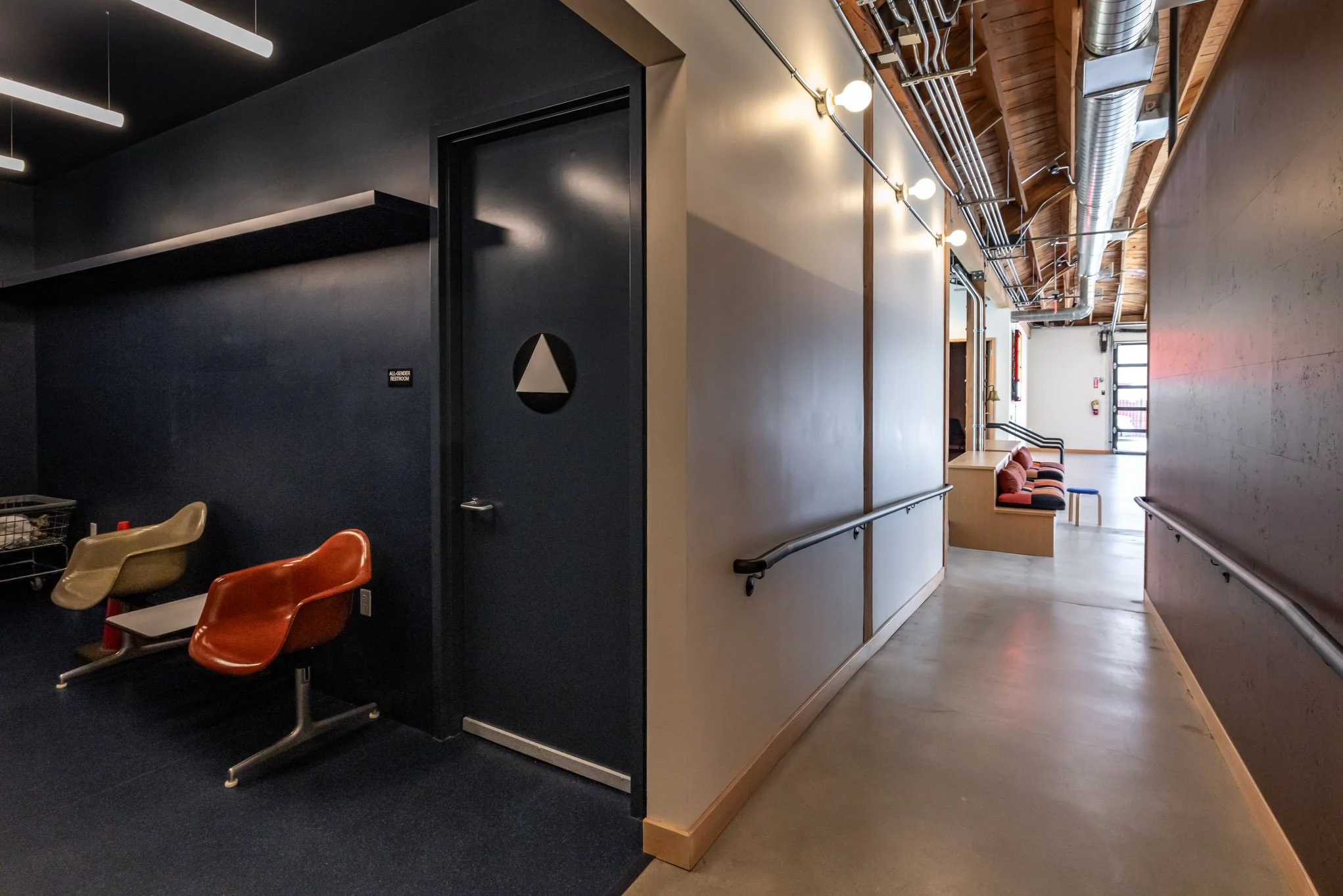 Interior hallway of a modern building with a dark-colored doorway, industrial ductwork, and seating area with orange and beige chairs.