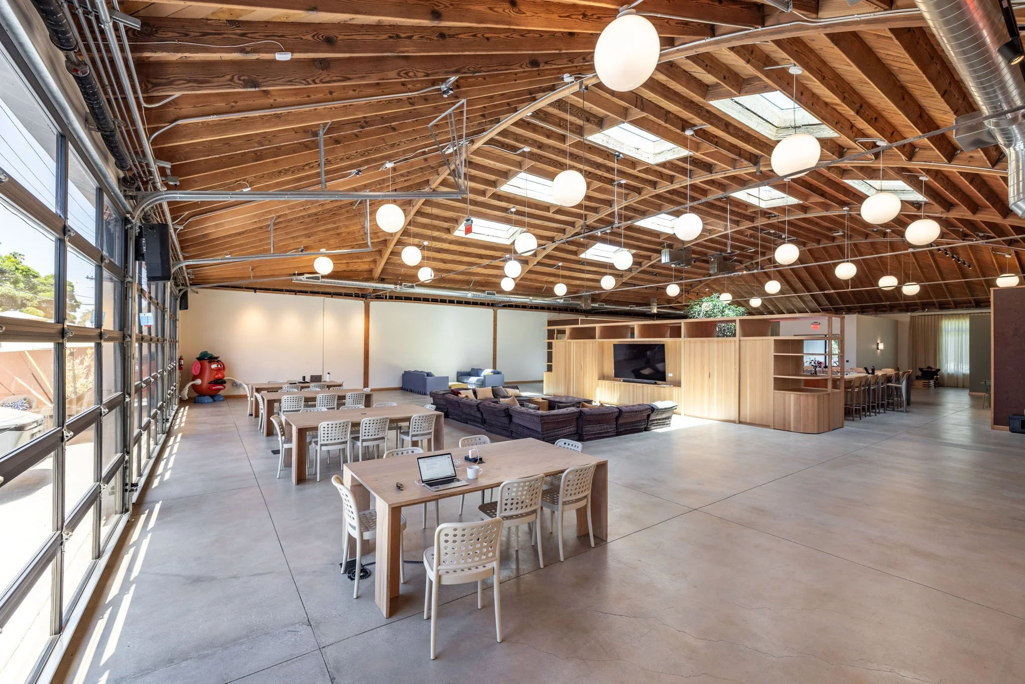 Spacious, modern event hall with wooden ceiling, large windows, and multiple round hanging lights, furnished with tables, chairs, a TV, and lounge seating.