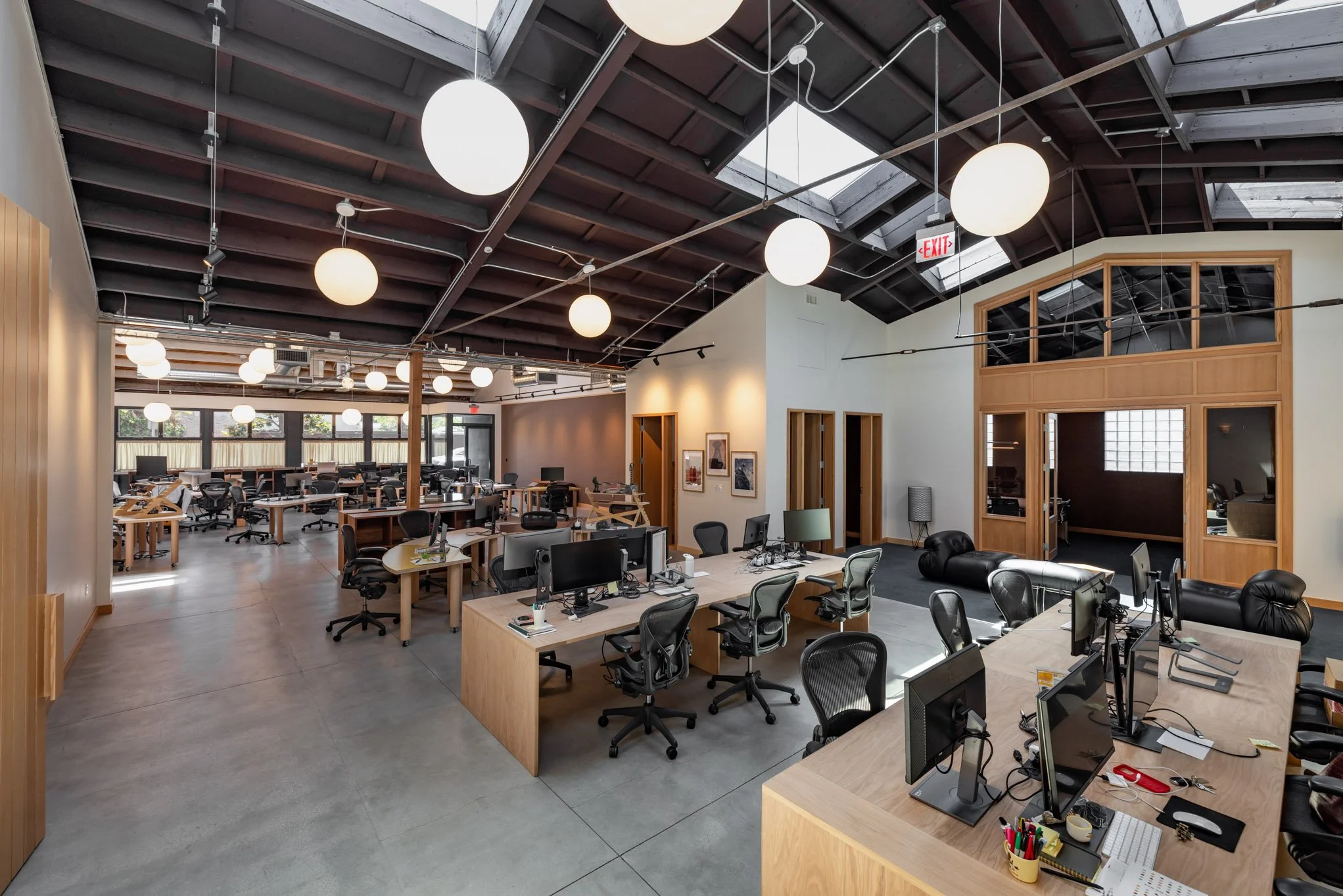 Open-plan modern office with multiple workstations, black office chairs, desktop computers, and wooden furniture. The ceiling is high with dark beams and round hanging lights. There are large windows bringing in natural light, and small rooms or offi