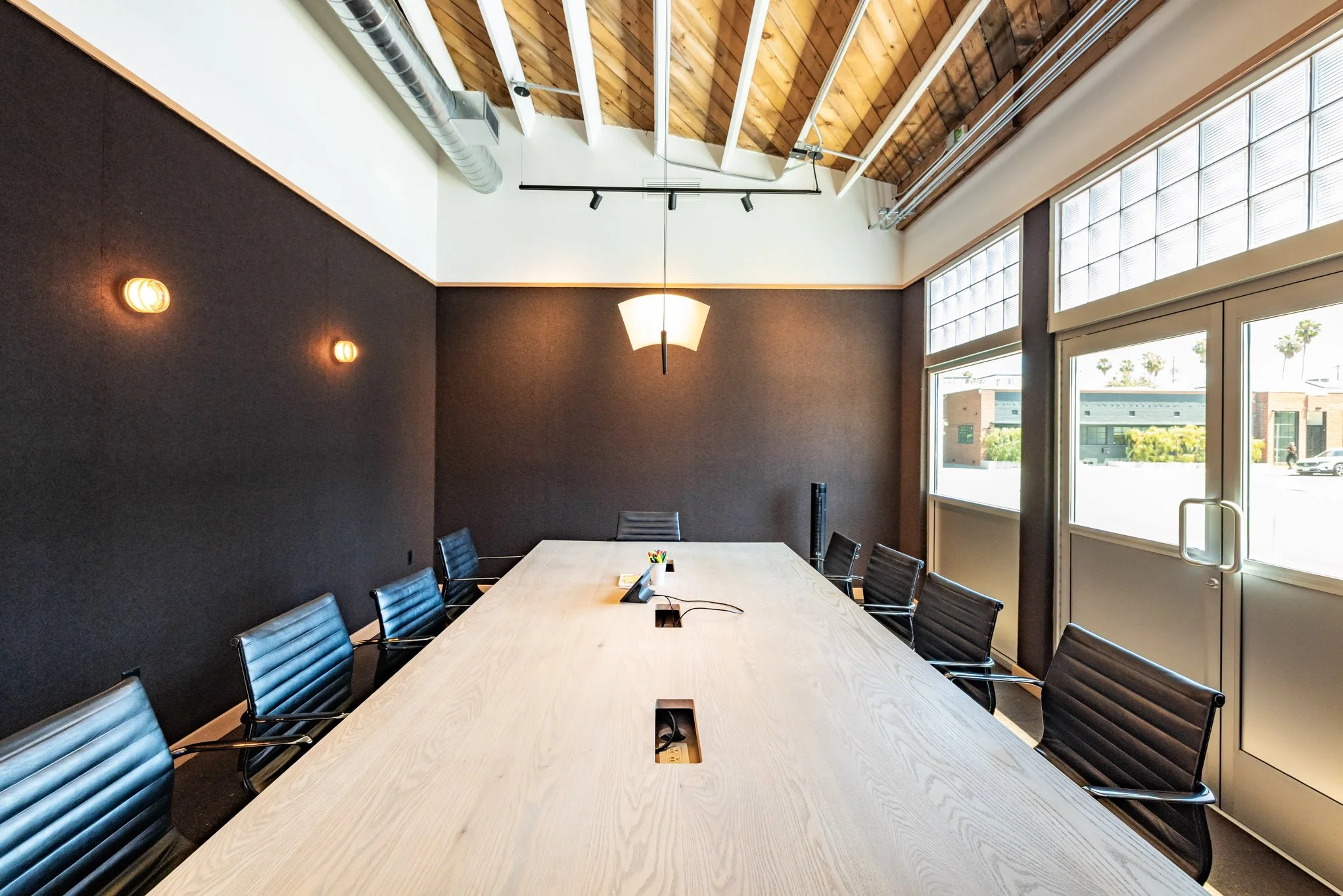 Empty modern conference room with a long wooden table, black leather chairs, large windows, and minimal lighting.