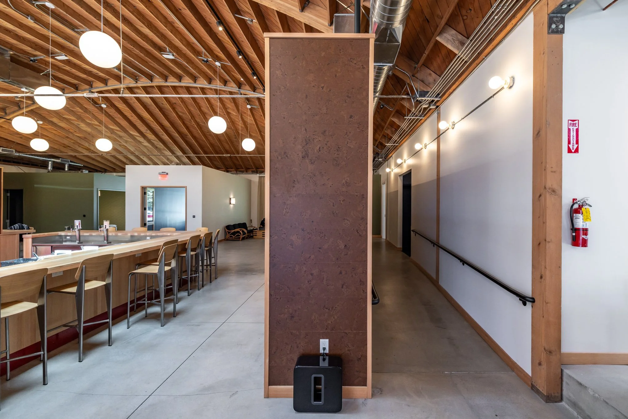 Interior of a modern building with a bar area on the left, a brown decorative wall in the middle, and a white wall with a handrail on the right. The ceiling is wooden with round pendant lights.
