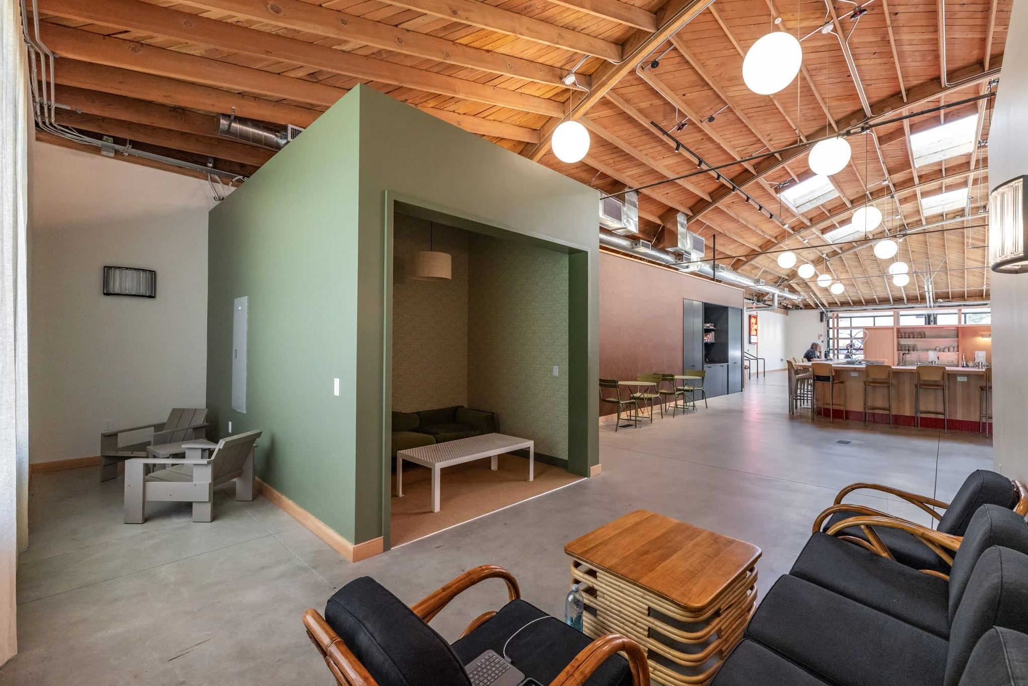 Interior of a spacious open-plan cafe or lounge with wooden ceiling beams, round hanging lights, a bar area with chairs, a seating nook with a sofa and a table, and additional chairs along the wall.