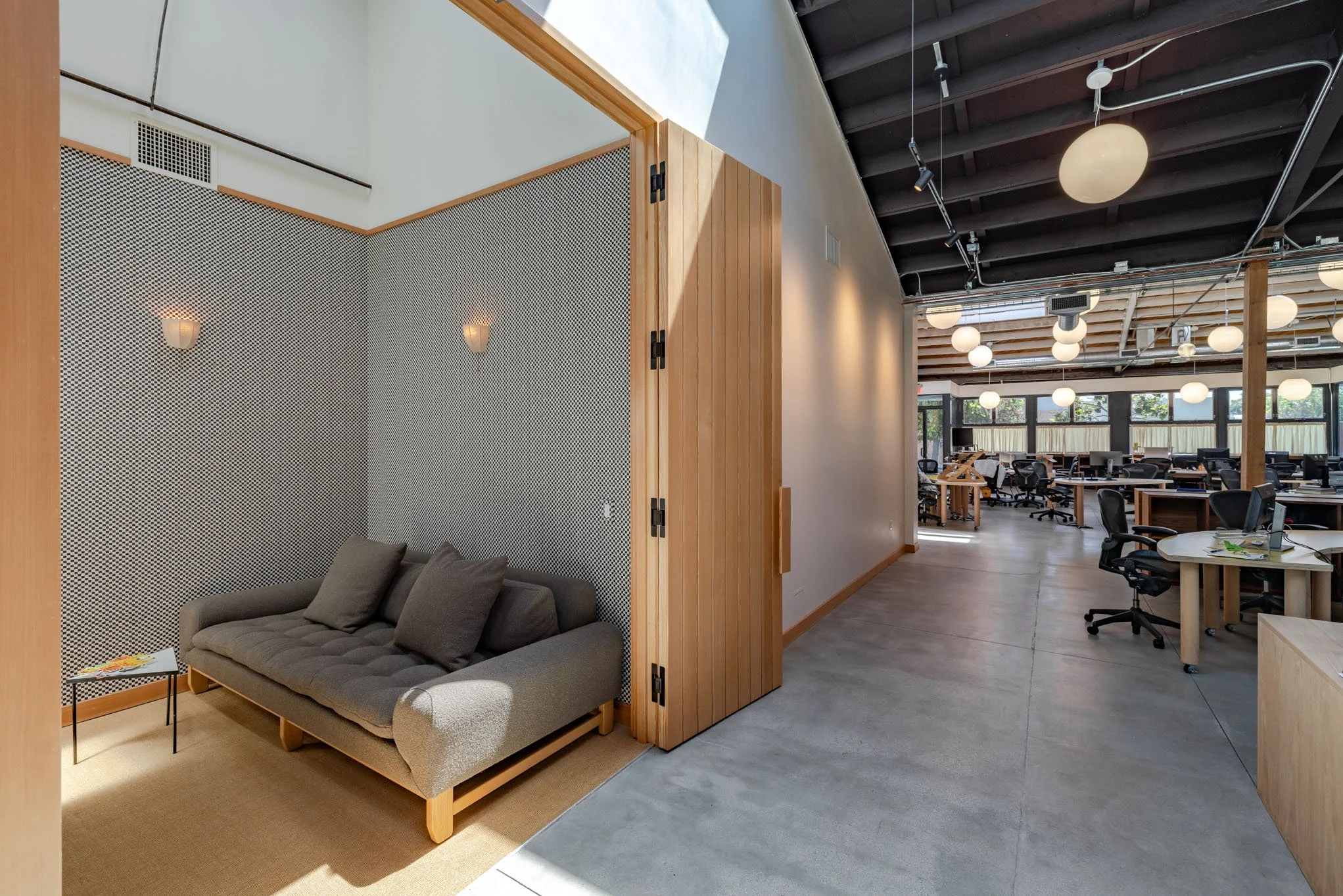 An office lounge area with a gray sofa and side table with magazines, adjacent to an open office workspace with multiple desks, chairs, and round ceiling lights.