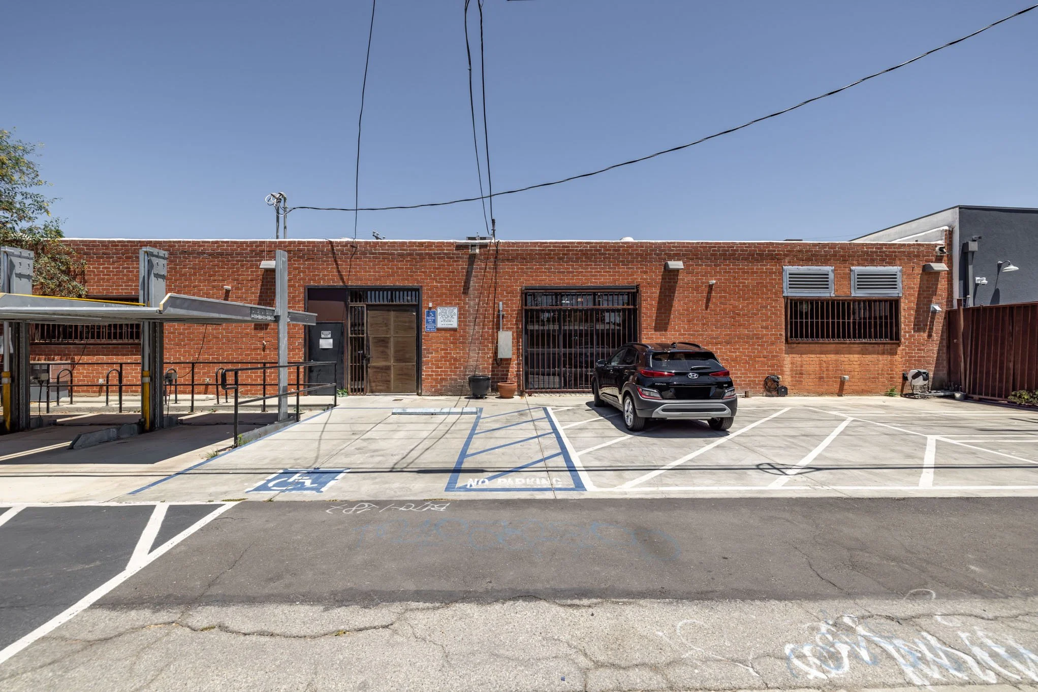 Parking lot with a single black car, designated disabled parking space, no parking signs, brick building with barred windows, and a clear blue sky.