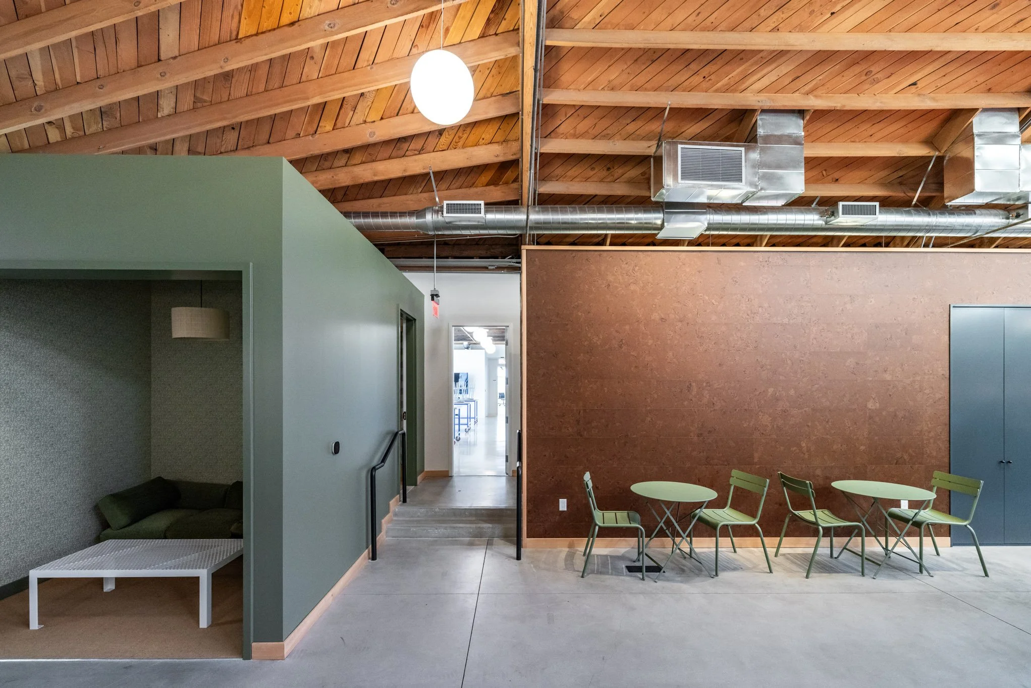 Interior of a modern space with a green partitioned nook with a gray sofa and white coffee table, four green chairs around two small green tables, and a wooden ceiling with exposed ducts and lighting.