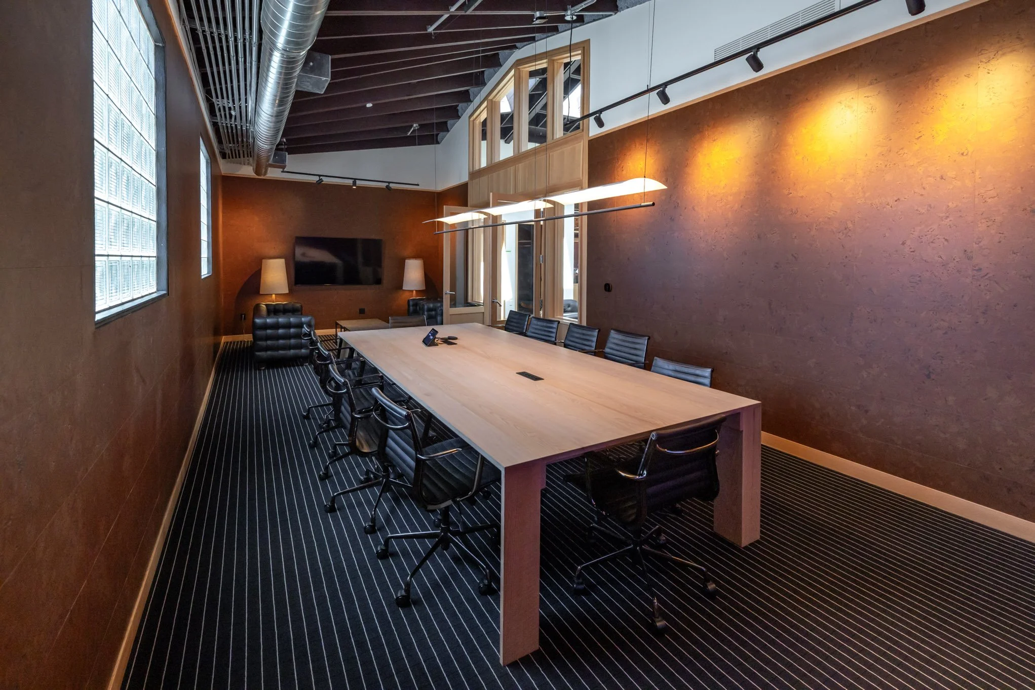 A modern conference room with a large wooden table, black office chairs, and a television on the back wall, with brown textured walls and track lighting.