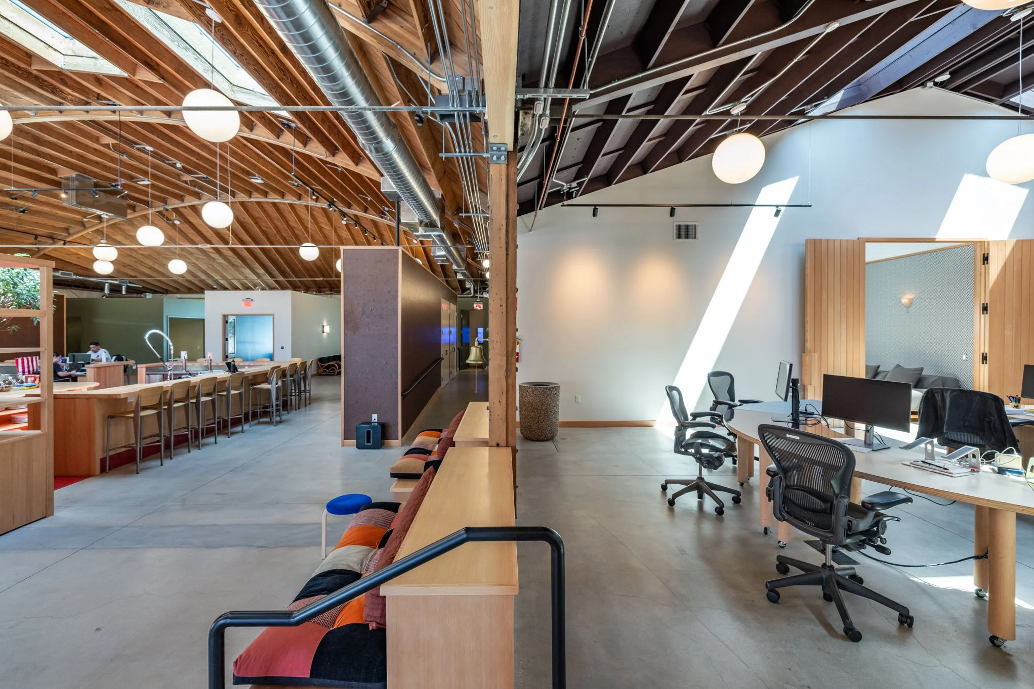 modern open-plan office behind divided workspace with desks, chairs, and computers, featuring a high wooden ceiling with exposed beams and white spherical hanging lights