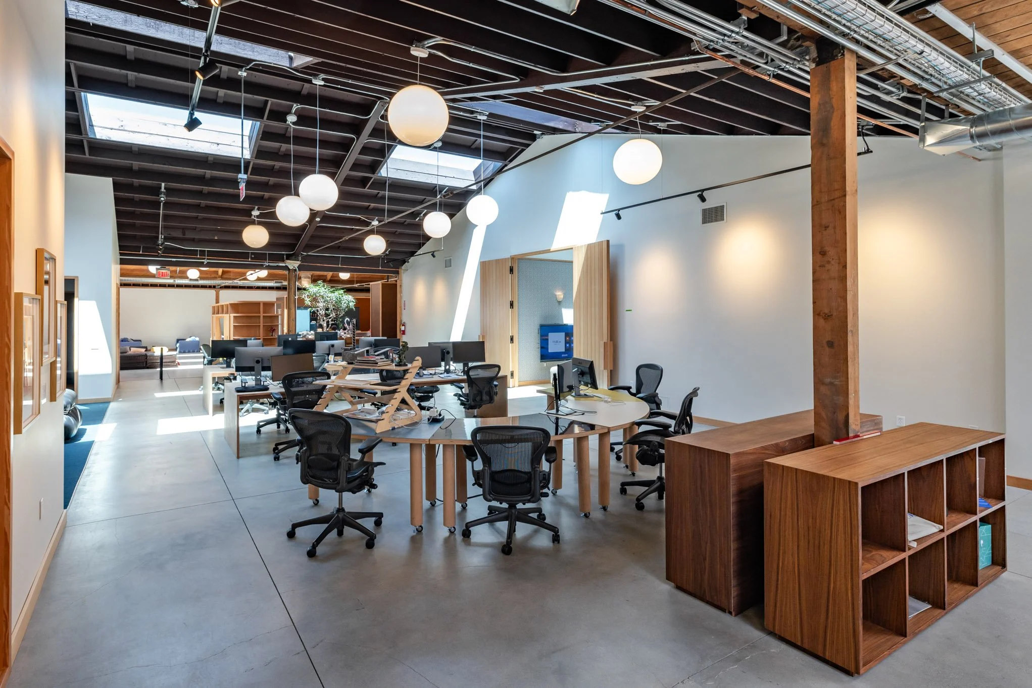 Open office space with modern design, multiple desks and chairs, hanging spherical lights, and natural light from skylights.