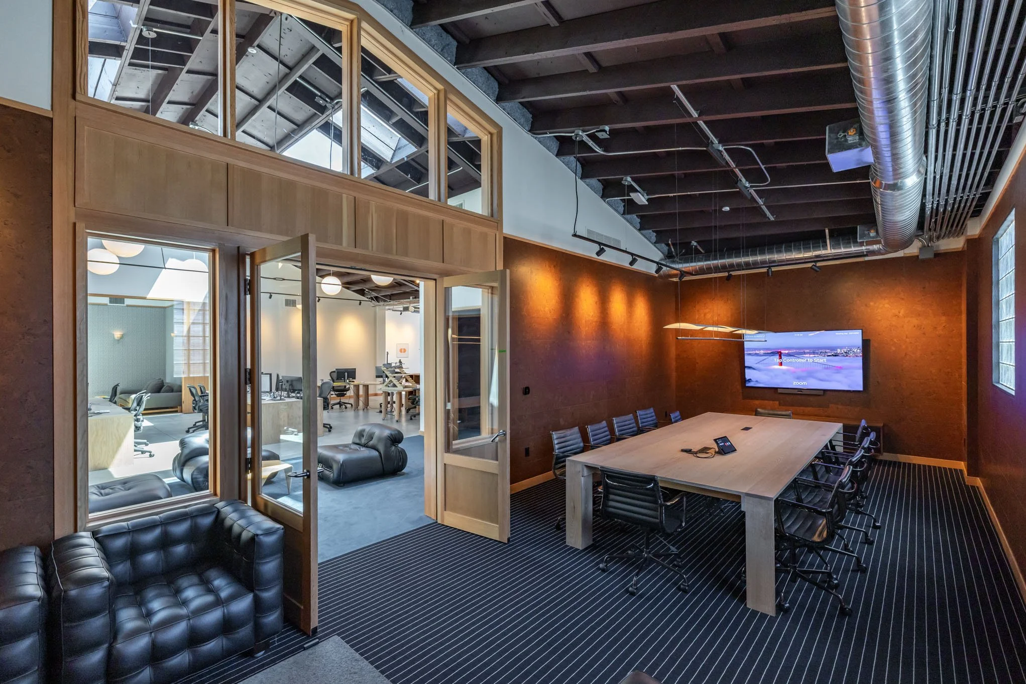 Conference room with a large wooden table and black chairs, a wall-mounted television, and an adjacent casual seating area with black leather chairs. Modern office space with exposed ceiling pipes, wooden accents, and large windows.