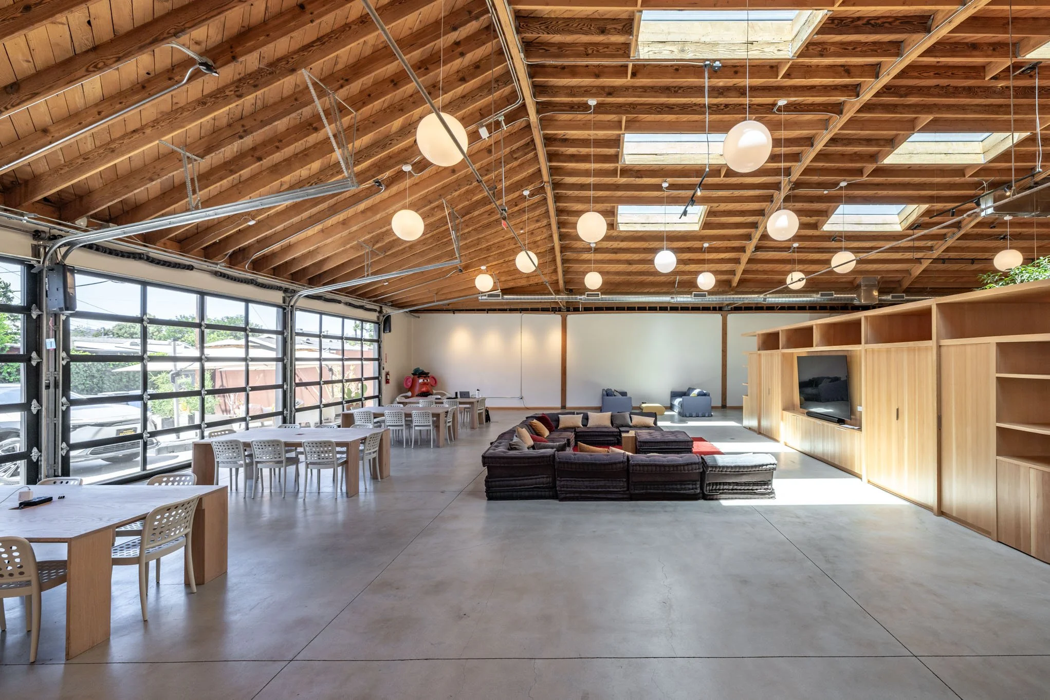 Spacious, modern lounge and meeting area with high wooden ceiling, large windows, tables with white chairs, a large sectional sofa with cushions, a smaller sofa, and a large wooden entertainment center with a TV.