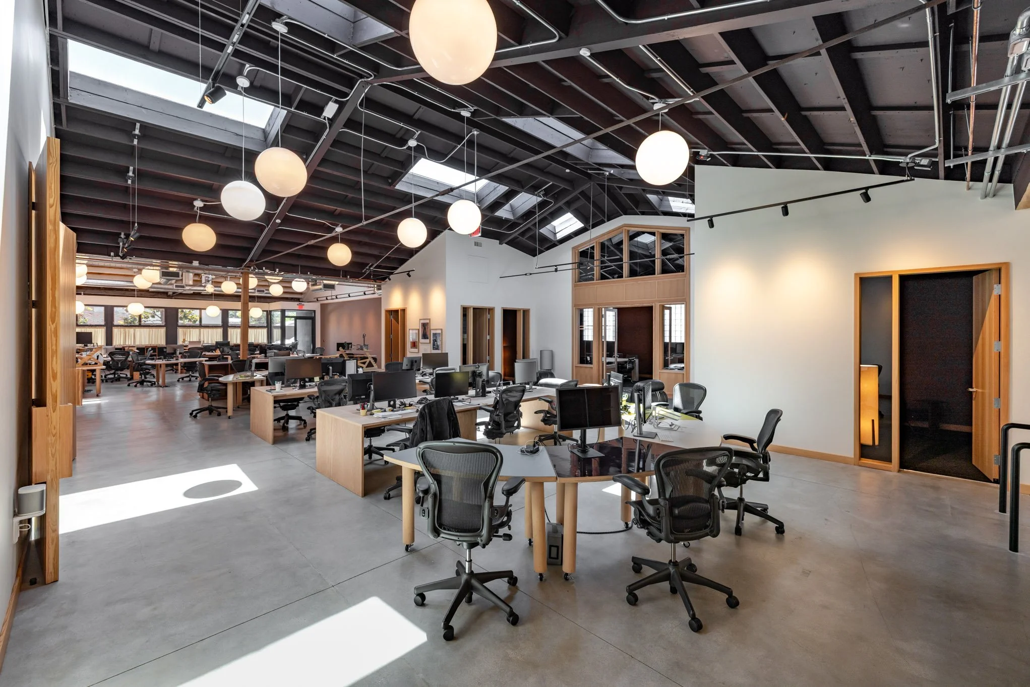 Modern open-plan office with multiple desks and ergonomic chairs, large windows, skylights, and hanging globe lights with an industrial ceiling.