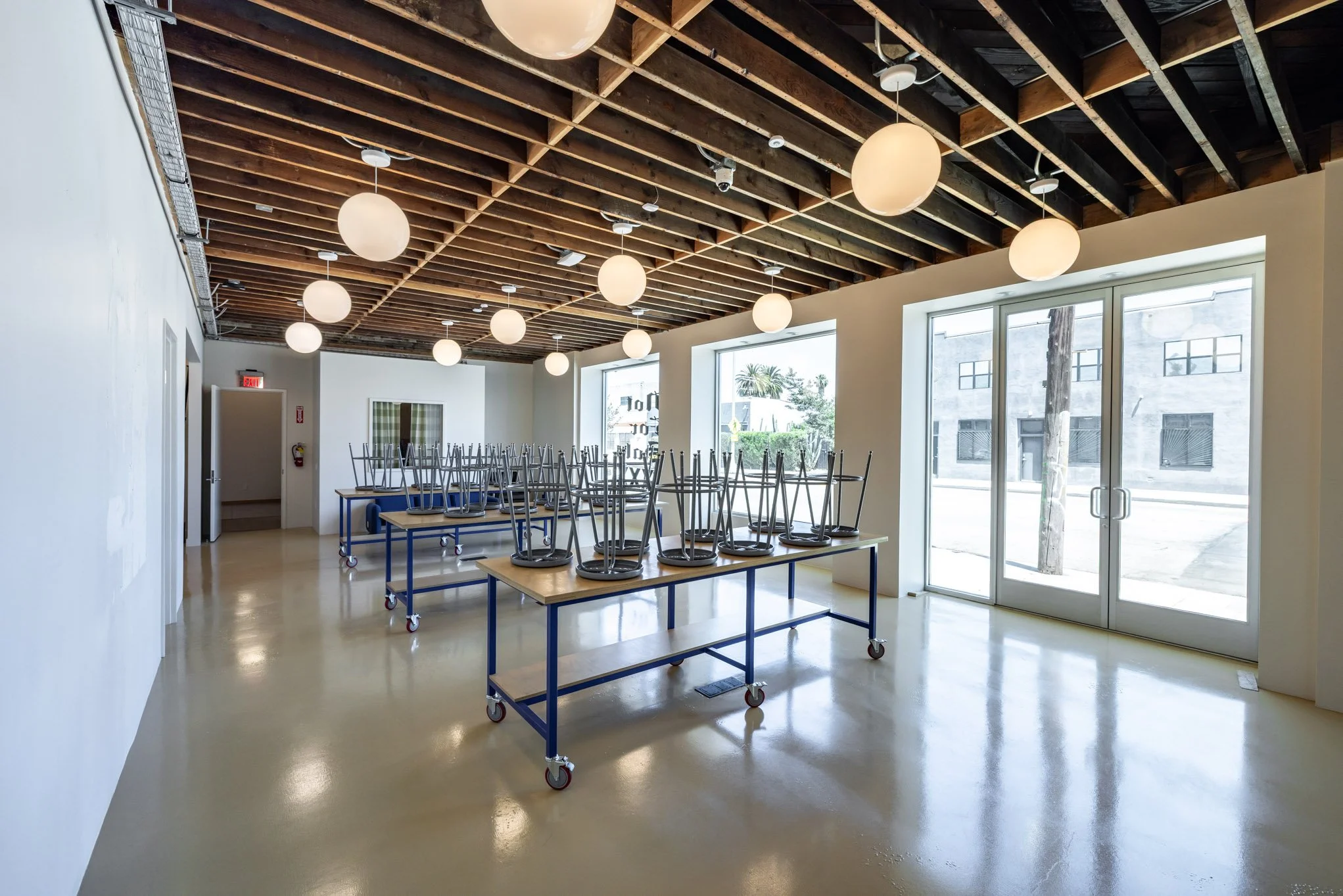Empty room with large windows, ceiling lights, and tables with stacked chairs on top, ready for setup.