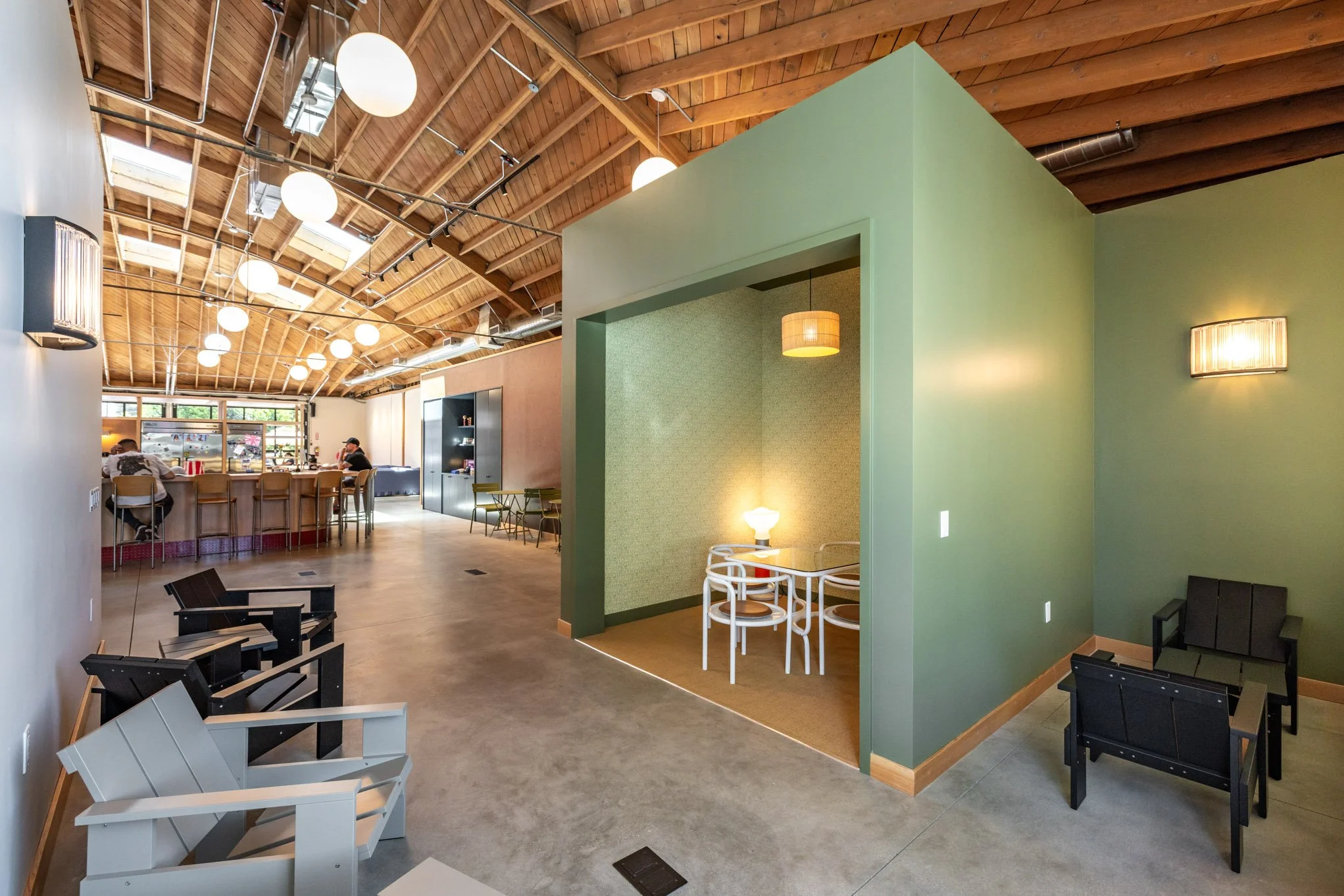 Interior view of a modern cafe with a high wooden ceiling, green and beige walls, and various seating areas, including armchairs, dining tables, and a barista station in the background.