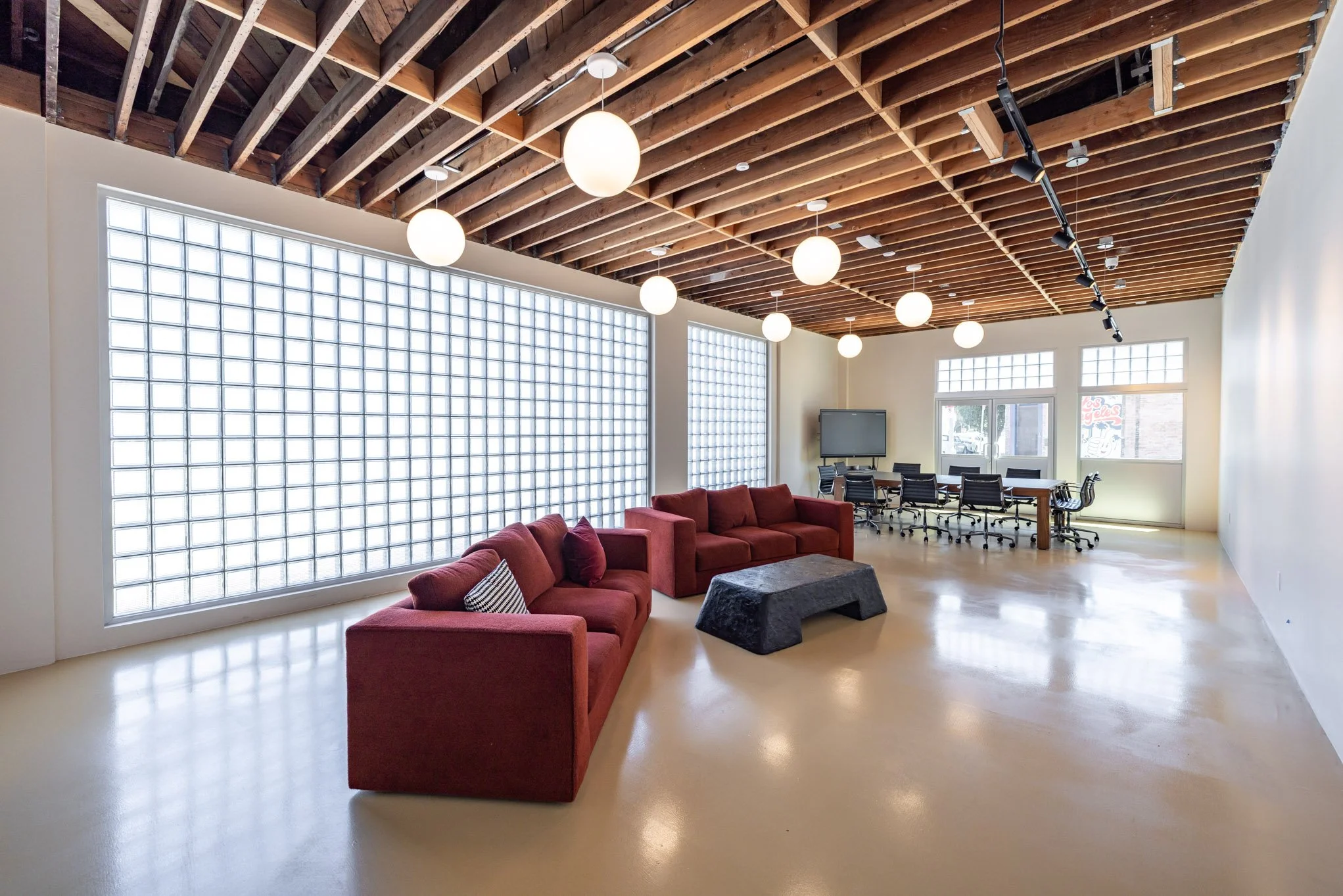 Modern open-concept office lounge with red couches, a black ottoman, a large glass block wall, and a communal table with chairs, illuminated by round pendant lights and track lighting, with wood ceiling beams.