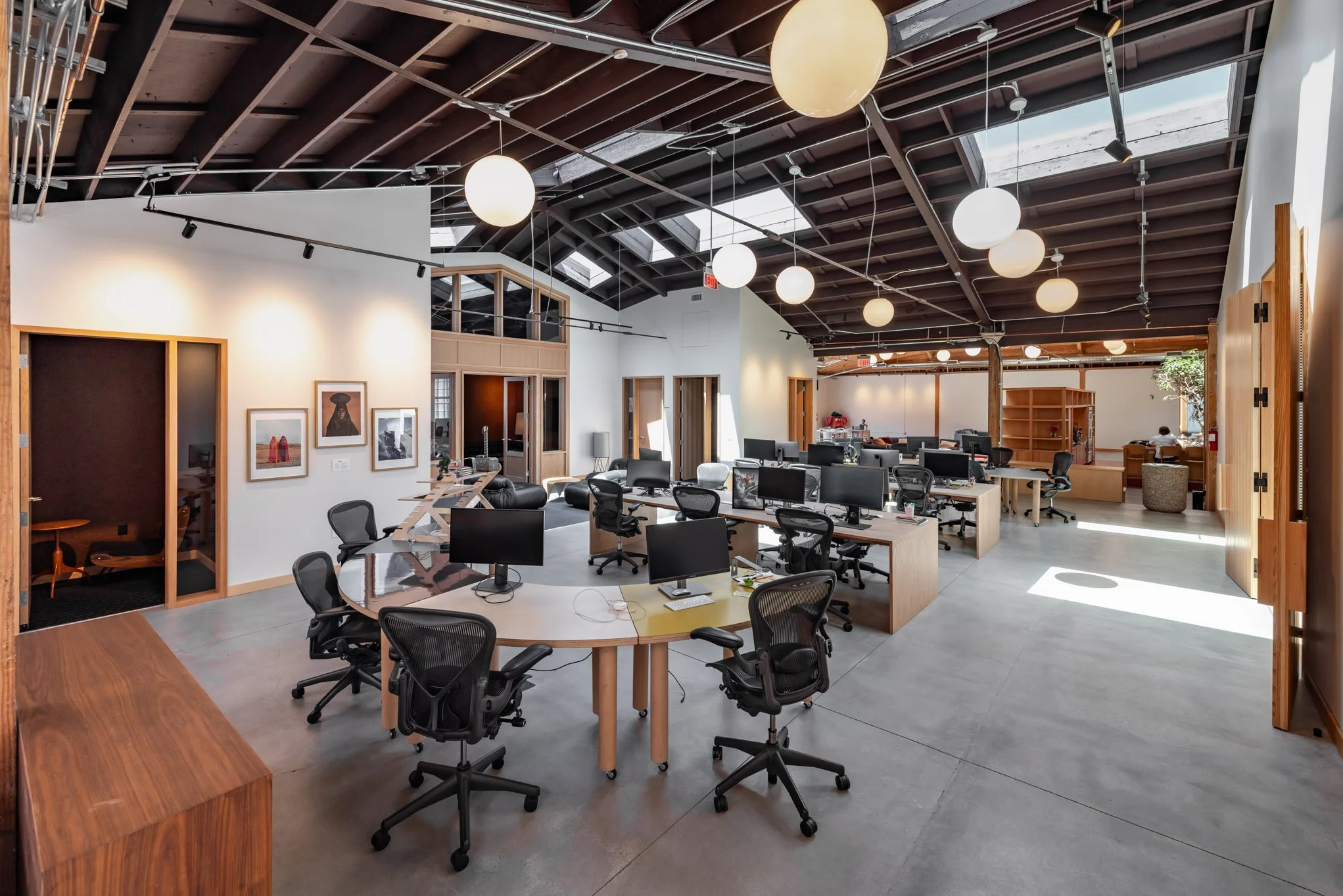 Modern open-plan office with multiple desks and ergonomic chairs, computer monitors, art on the wall, and large windows letting in natural light.
