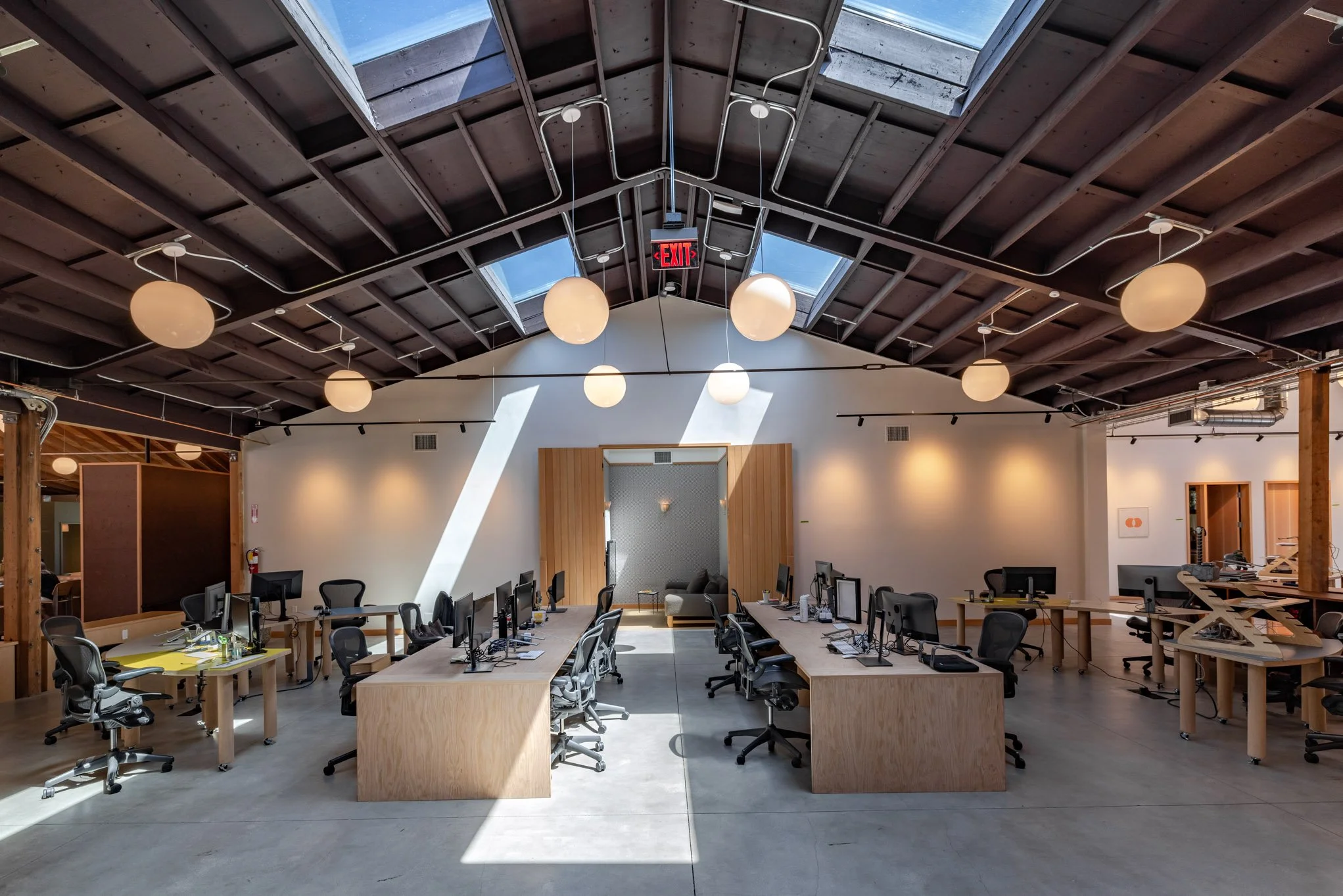 An open-concept modern office space with high ceilings, wooden beams, and skylights. Contains multiple workstations with black chairs and computer monitors, and a cozy seating area with a gray couch in the background.