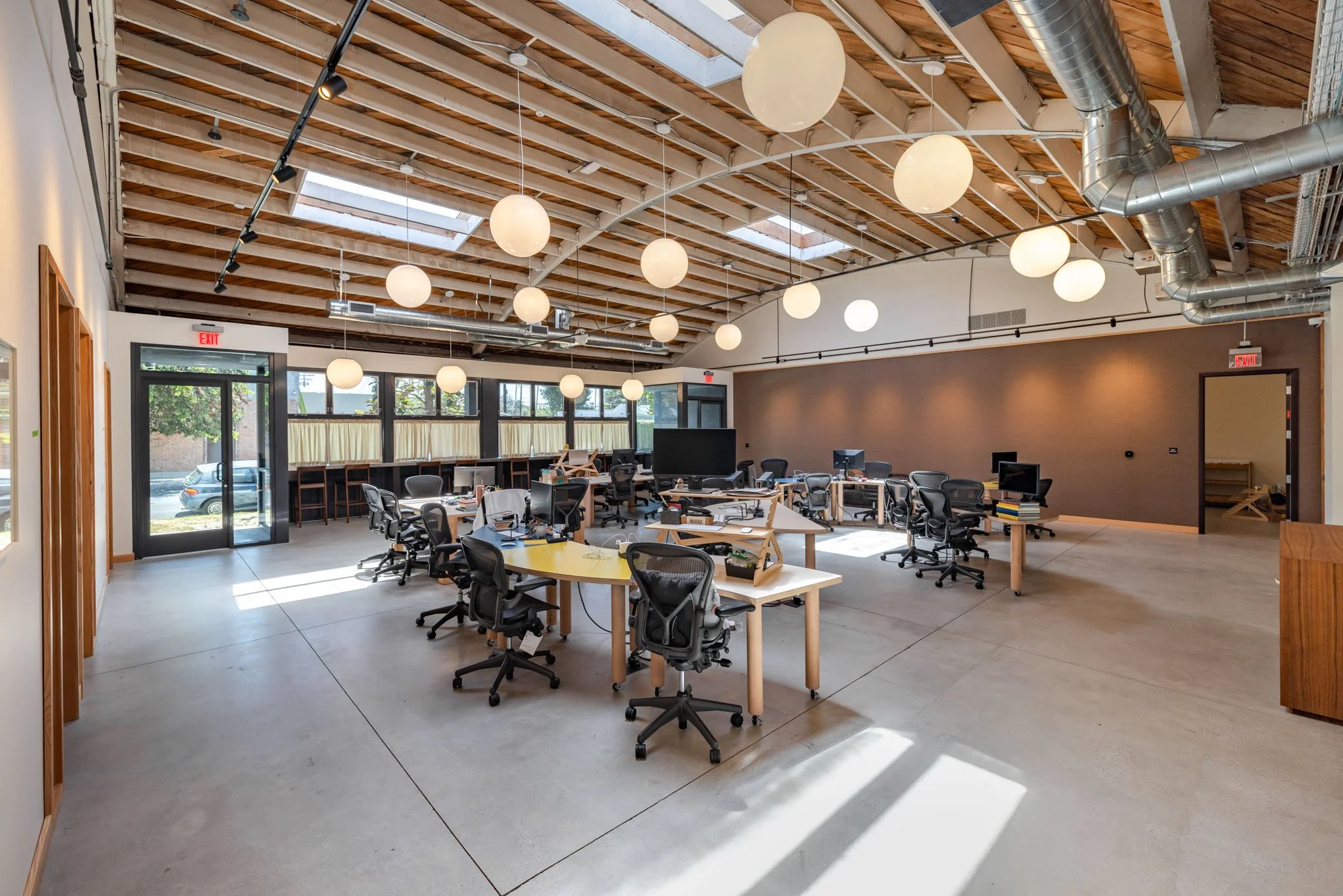 Empty modern office space with multiple desks, chairs, computers, and large windows, with ceiling lights and exposed ductwork.