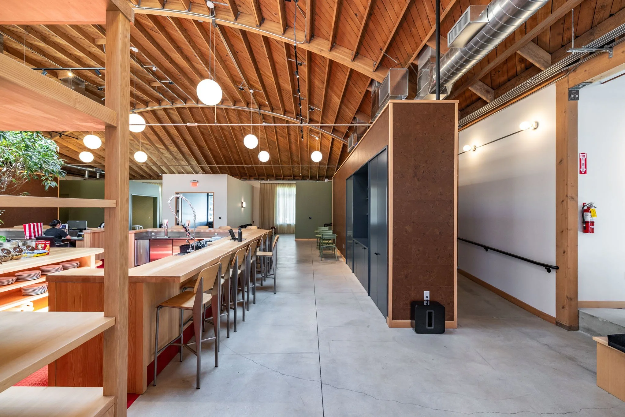 Interior of a modern, rustic-style cafe or dining area with wooden ceiling and furniture, a long bar counter with chairs, some equipment, and a few people working in the background.